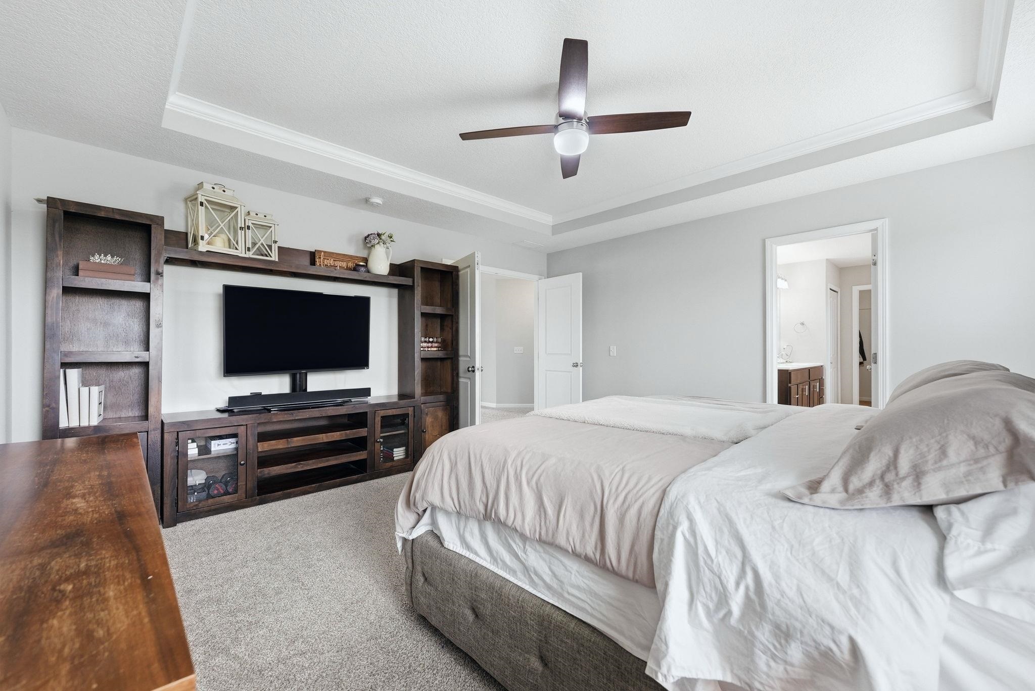Bedroom featuring a raised ceiling, crown molding, carpet flooring, ceiling fan, and ensuite bath