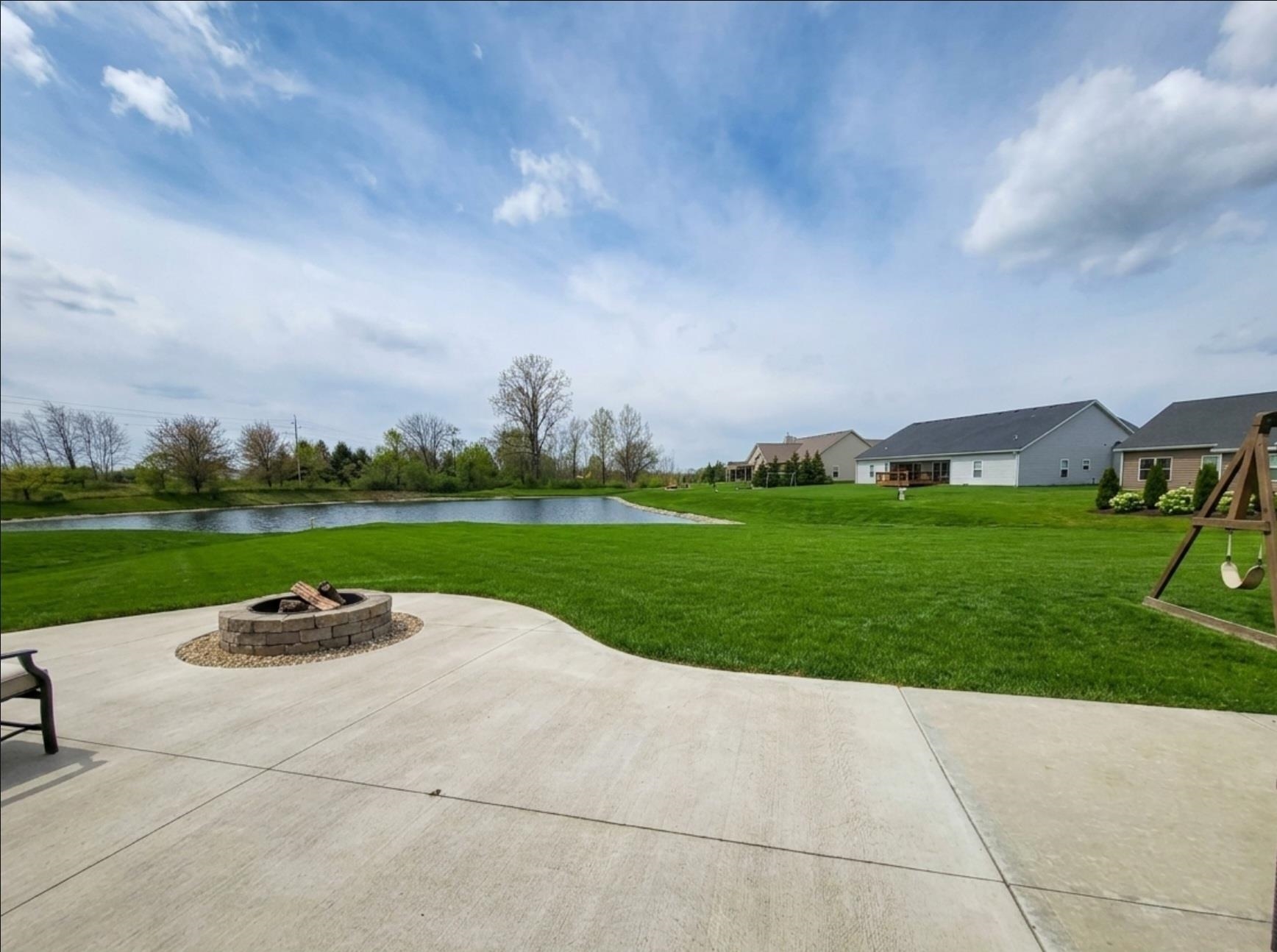 Expansive backyard featuring a curved concrete patio with an integrated stone-faced fire pit, a sprawling green lawn, and a serene pond