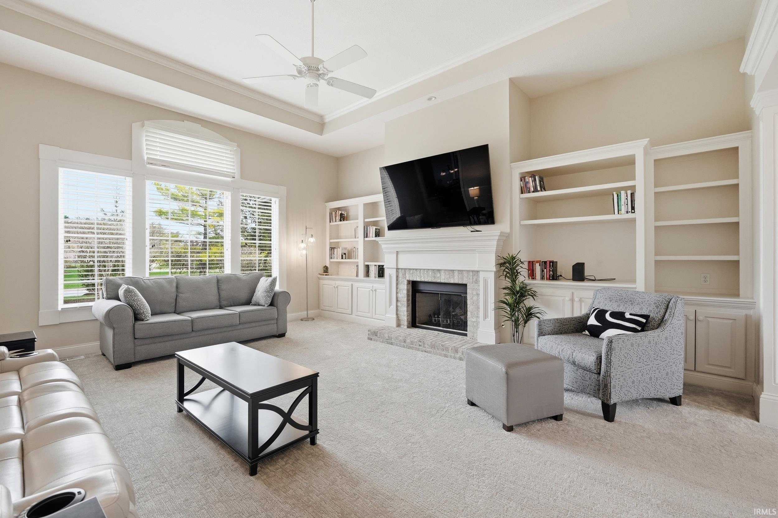 Living area featuring a raised ceiling, a ceiling fan, a brick fireplace, light colored carpet, and built in features