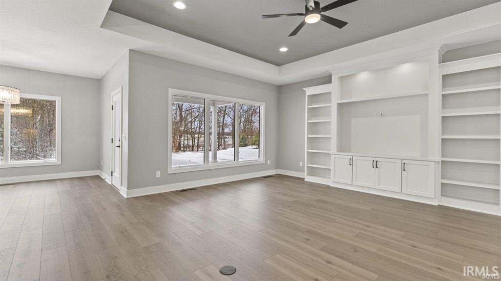 Unfurnished living room featuring a raised ceiling, a ceiling fan, light wood-type flooring, built in features, and a chandelier