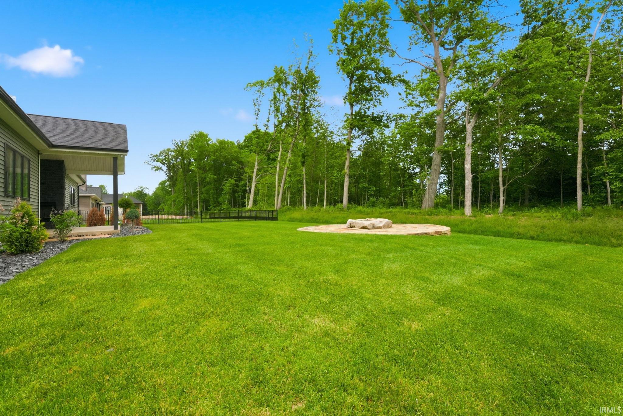 Expansive lawn area featuring a stone fire pit with stone surround, mature trees bordering the property, and a covered porch with black columns