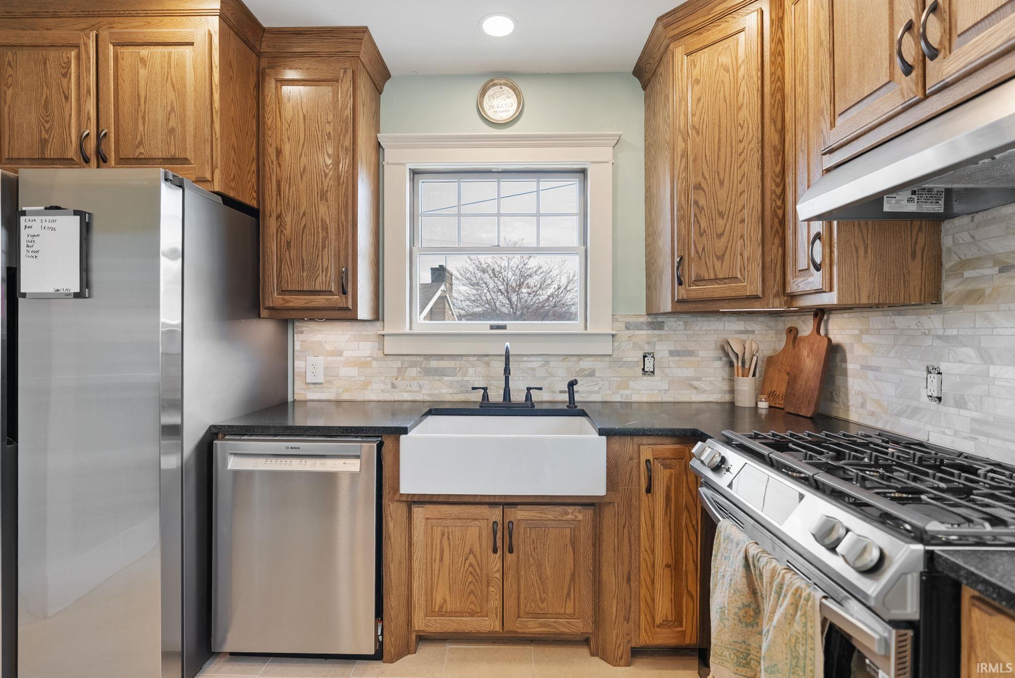 Kitchen featuring stainless steel appliances, wood finish cabinetry, dark stone counters, light tile patterned flooring, and decorative backsplash