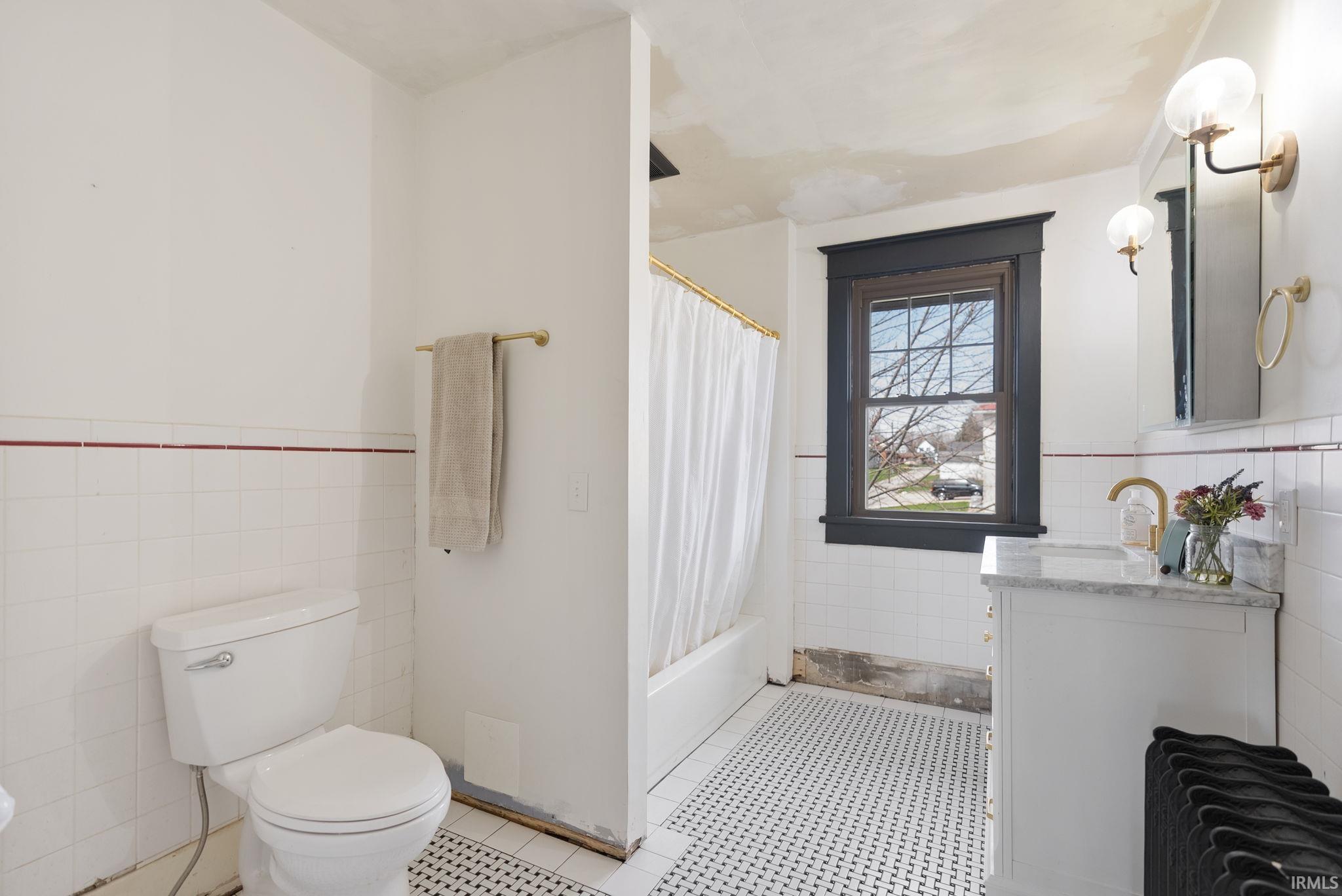 Bathroom featuring tile walls, vanity, a wainscoted wall, shower / bathtub combination with curtain, and light tile patterned floors