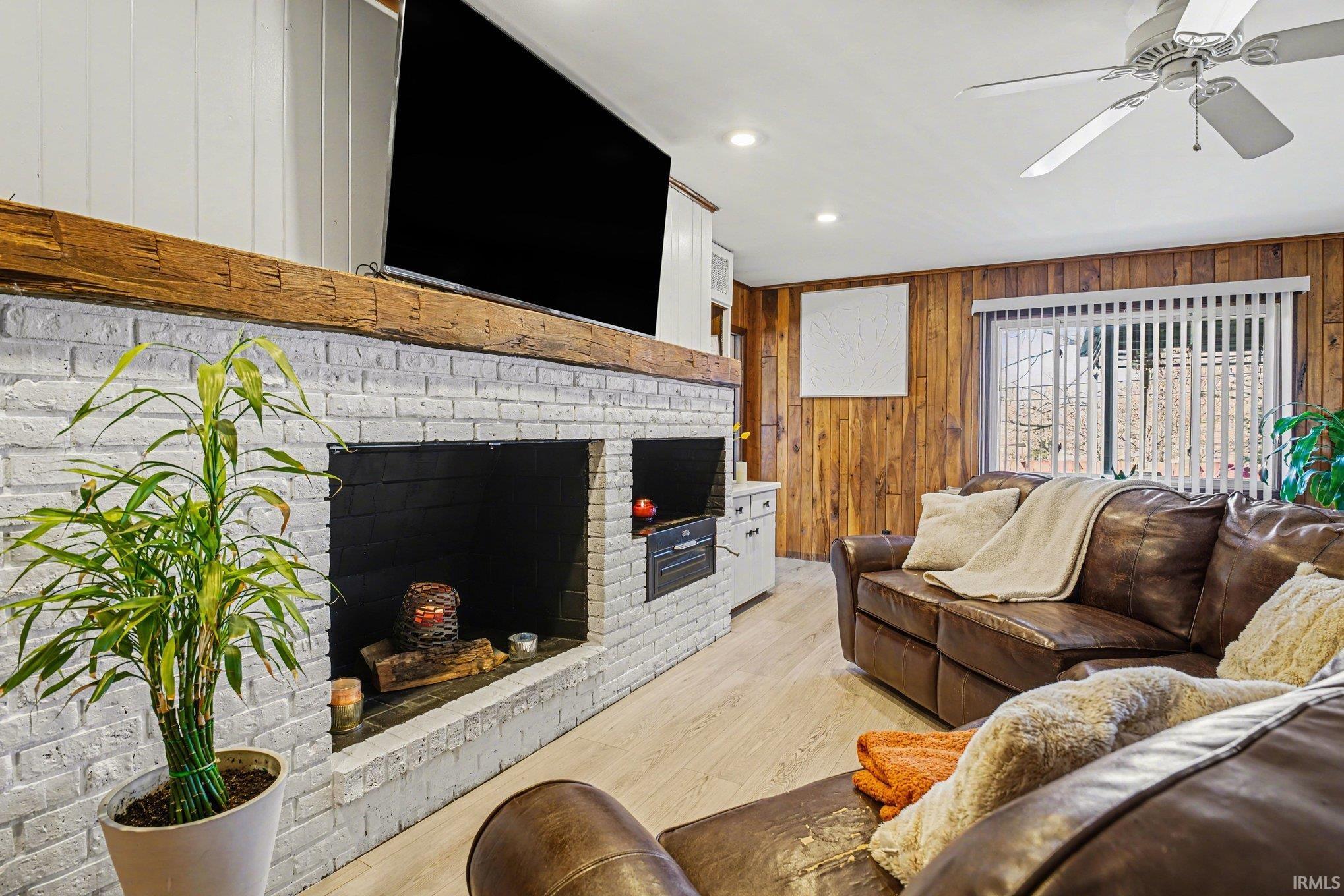 Living room with wood walls, light wood-type flooring, a ceiling fan, a brick fireplace, and recessed lighting