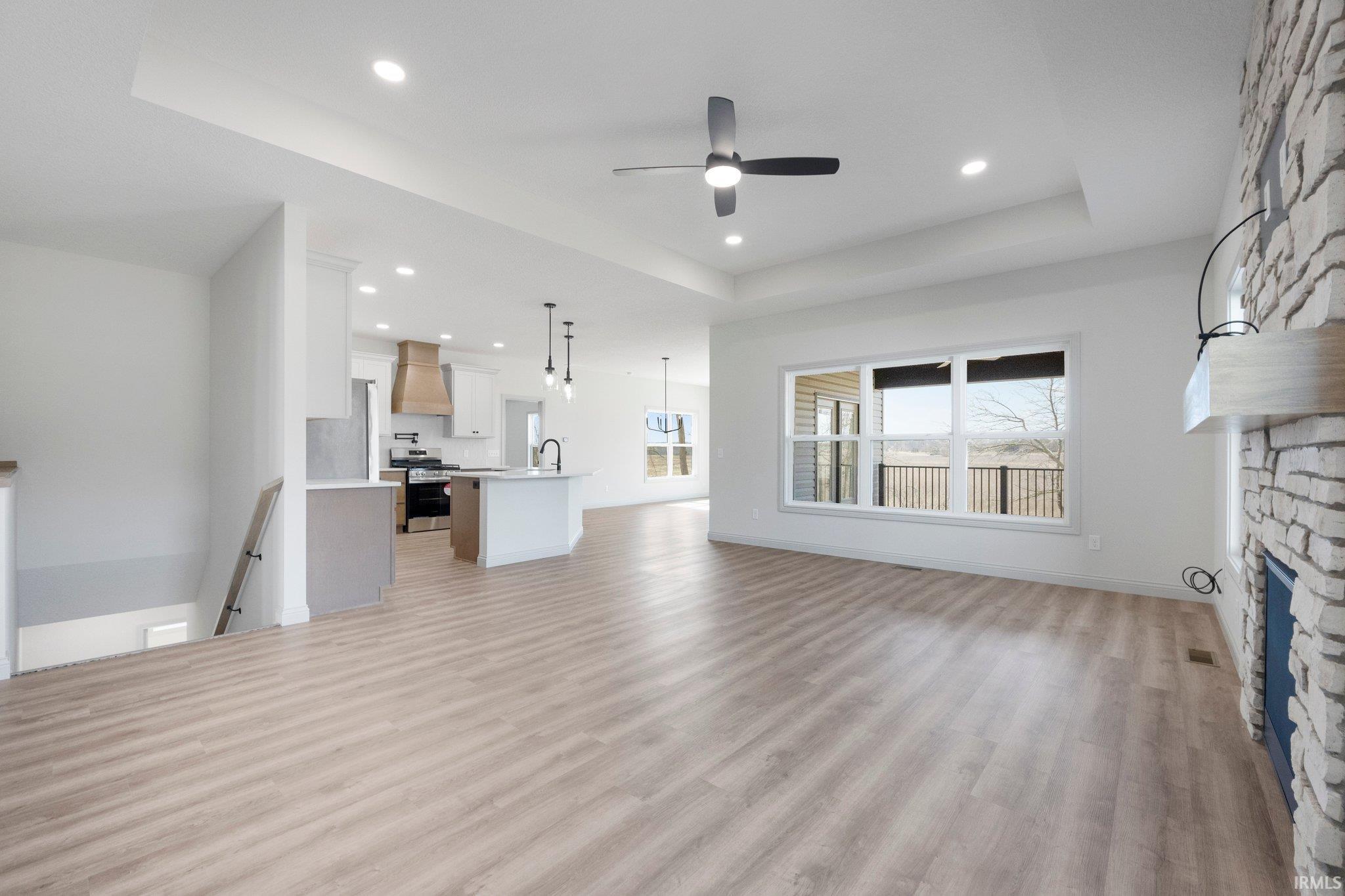 Open concept living area featuring light wood-finish flooring, a stone fireplace with a wood mantle, and a tray ceiling with recessed lighting and a ceiling fan