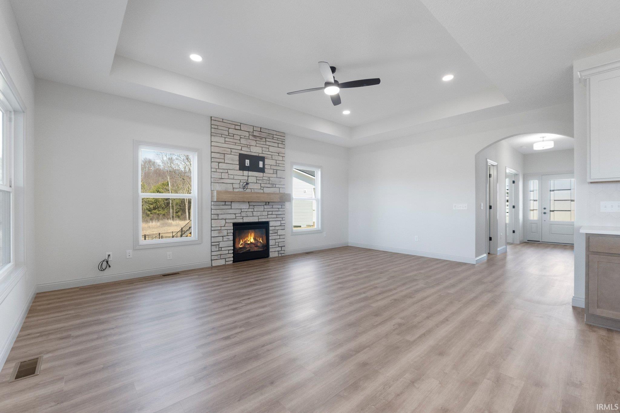 Spacious living area featuring a stone-faced fireplace with a wood mantel, light wood-finish flooring, a tray ceiling with recessed lighting, and a ceiling fan