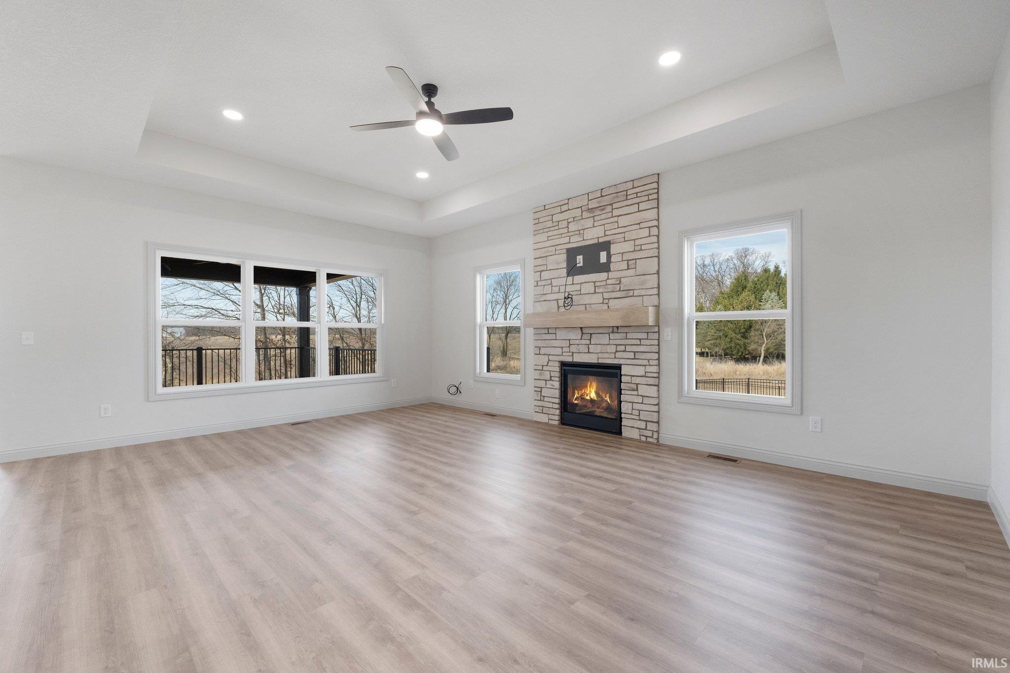Spacious room featuring wood-finish flooring, a stone-clad fireplace with a wood mantel, a tray ceiling with recessed lighting and a ceiling fan, and multiple windows offering outdoor scenery
