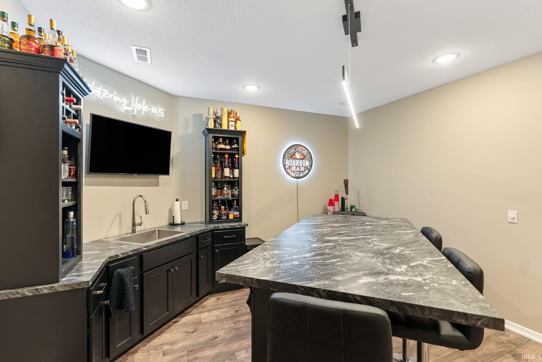 Indoor wet bar with dark countertops, light wood-style floors, dark cabinets, recessed lighting, and a textured ceiling