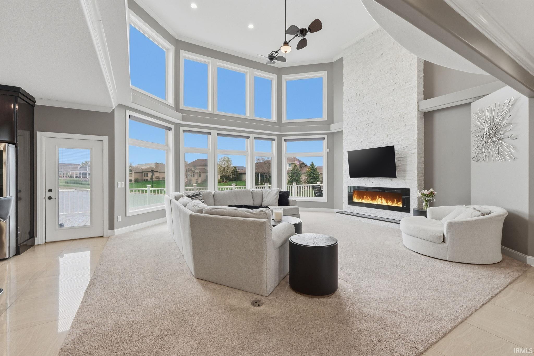 Living room featuring ceiling fan, a stone fireplace, light tile patterned floors, a high ceiling, and crown molding