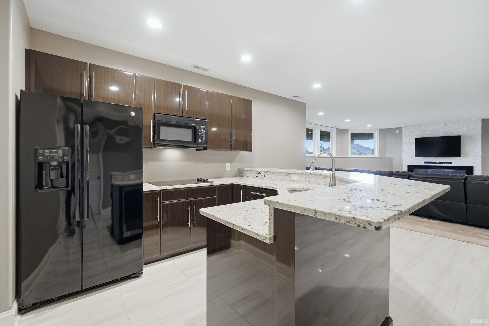 Kitchen featuring a peninsula, black appliances, dark wood finish cabinetry, open floor plan, and light stone countertops