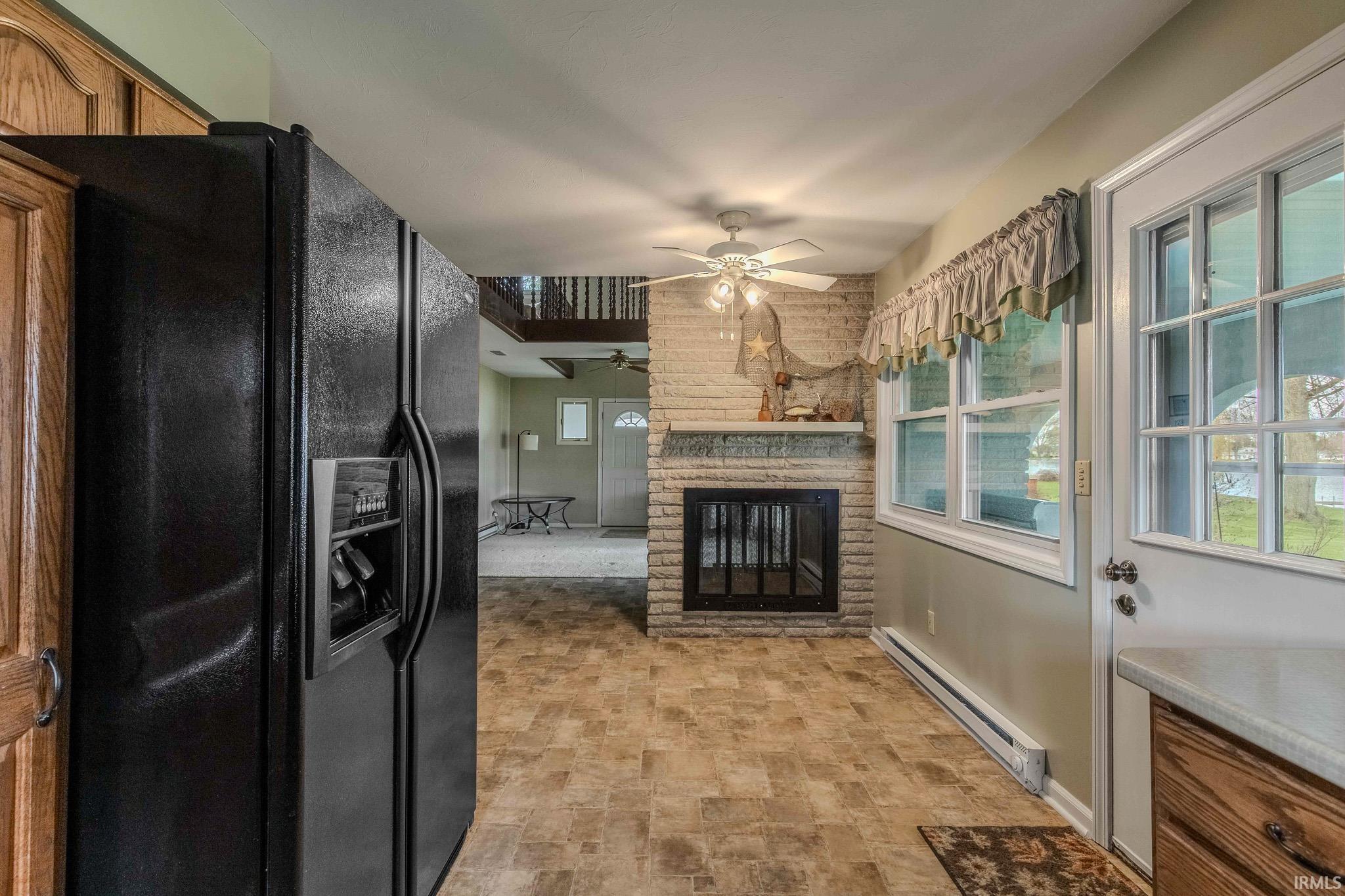 Kitchen with black fridge with ice dispenser, a baseboard heating unit, stone finish flooring, a brick fireplace, and ceiling fan