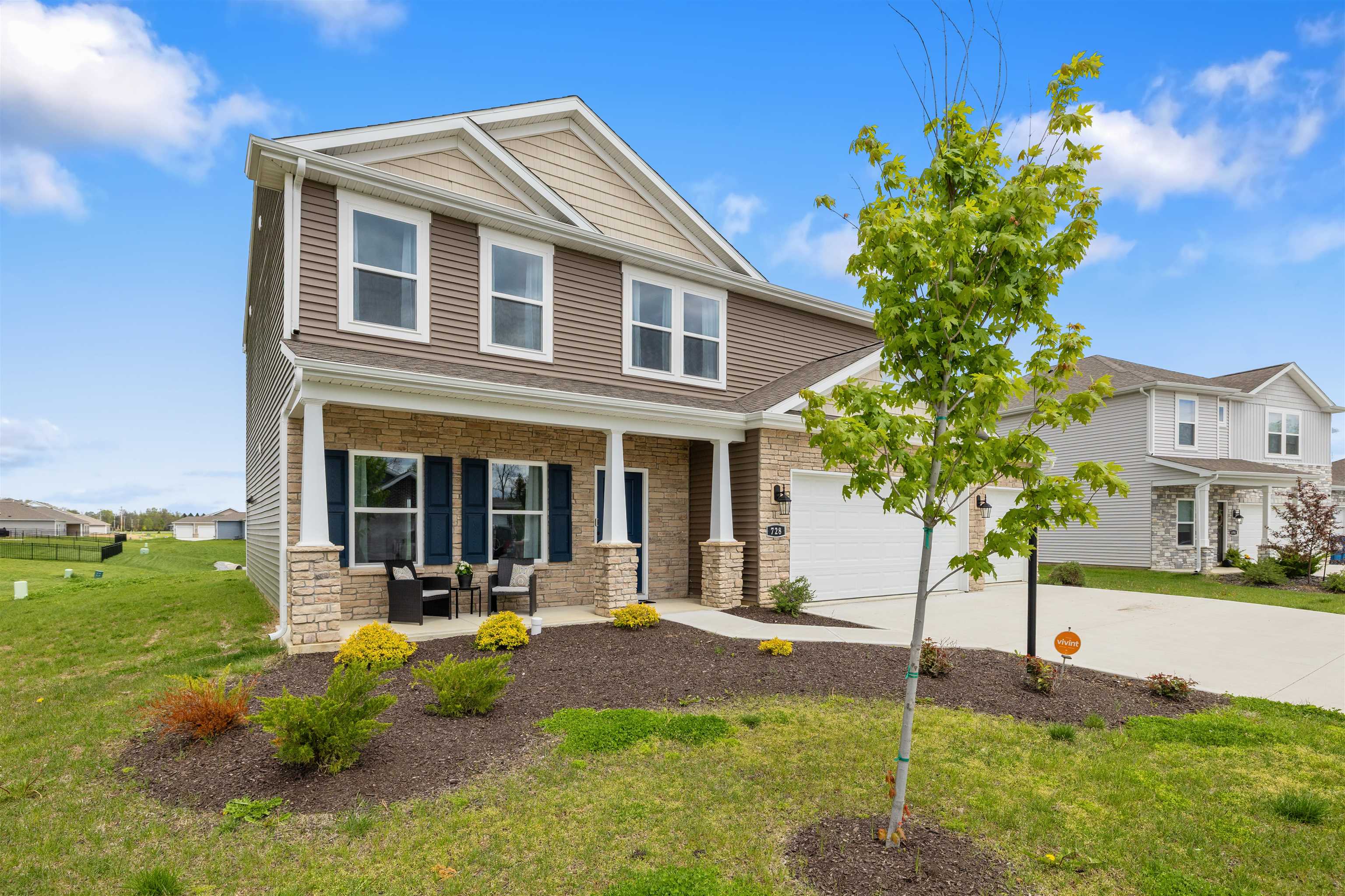 Two-story residential exterior featuring a stone and siding facade, a covered front porch, an attached garage, a paved driveway, and mature landscaping