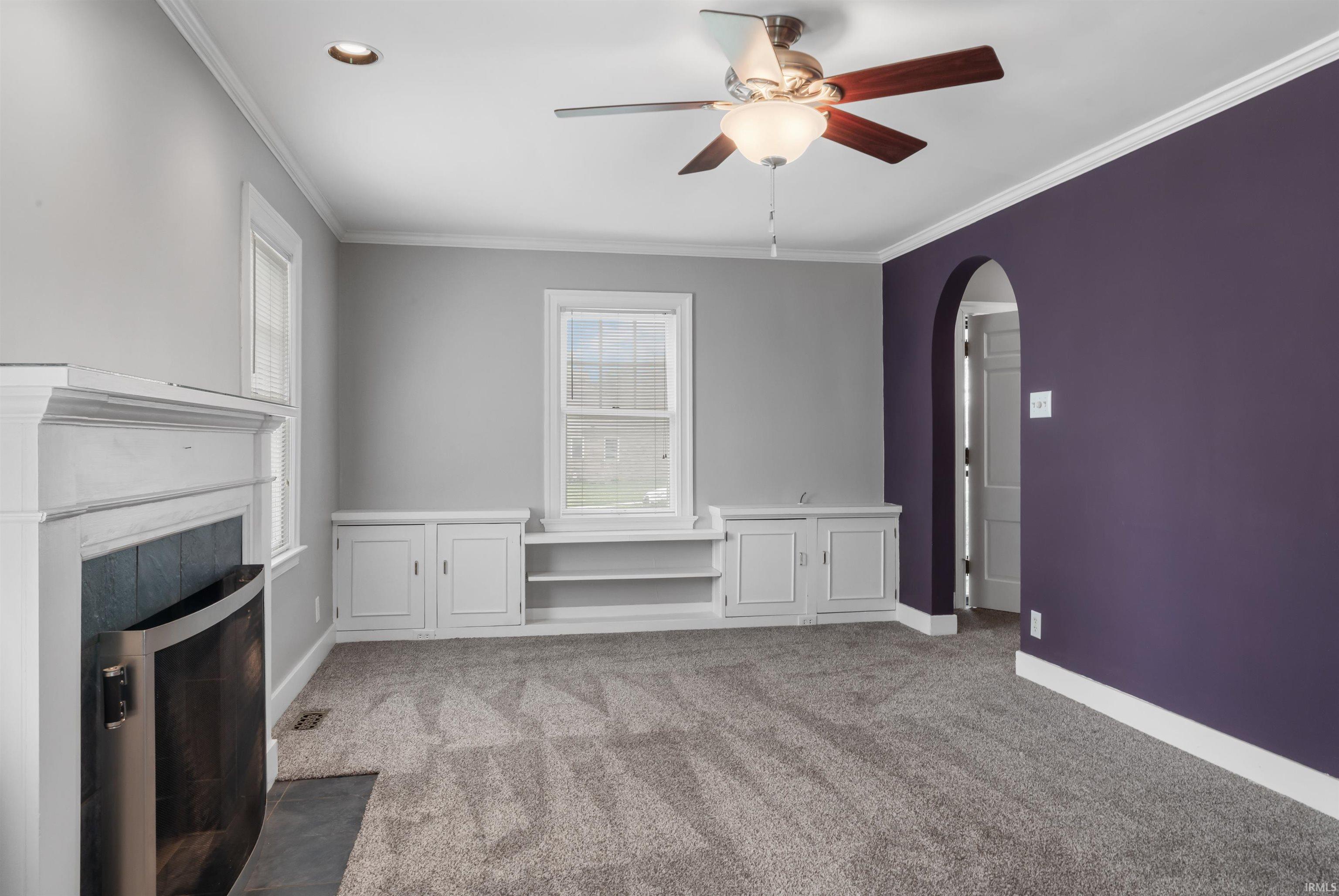 Unfurnished living room featuring arched walkways, a ceiling fan, dark carpet, crown molding, and a fireplace with flush hearth