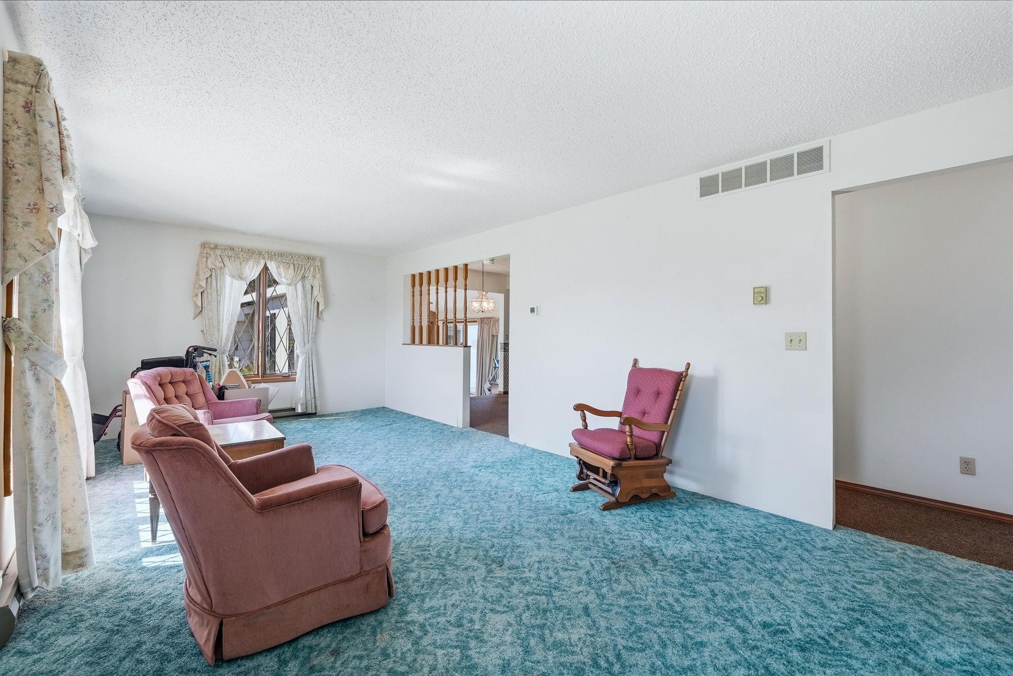 Living area featuring textured carpeting, white walls, and a large window with decorative trim