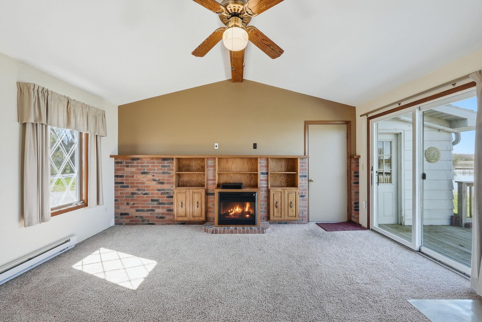 Living area featuring a brick-surround fireplace with built-in shelving, vaulted ceiling with a fan, and sliding glass door access to an exterior deck