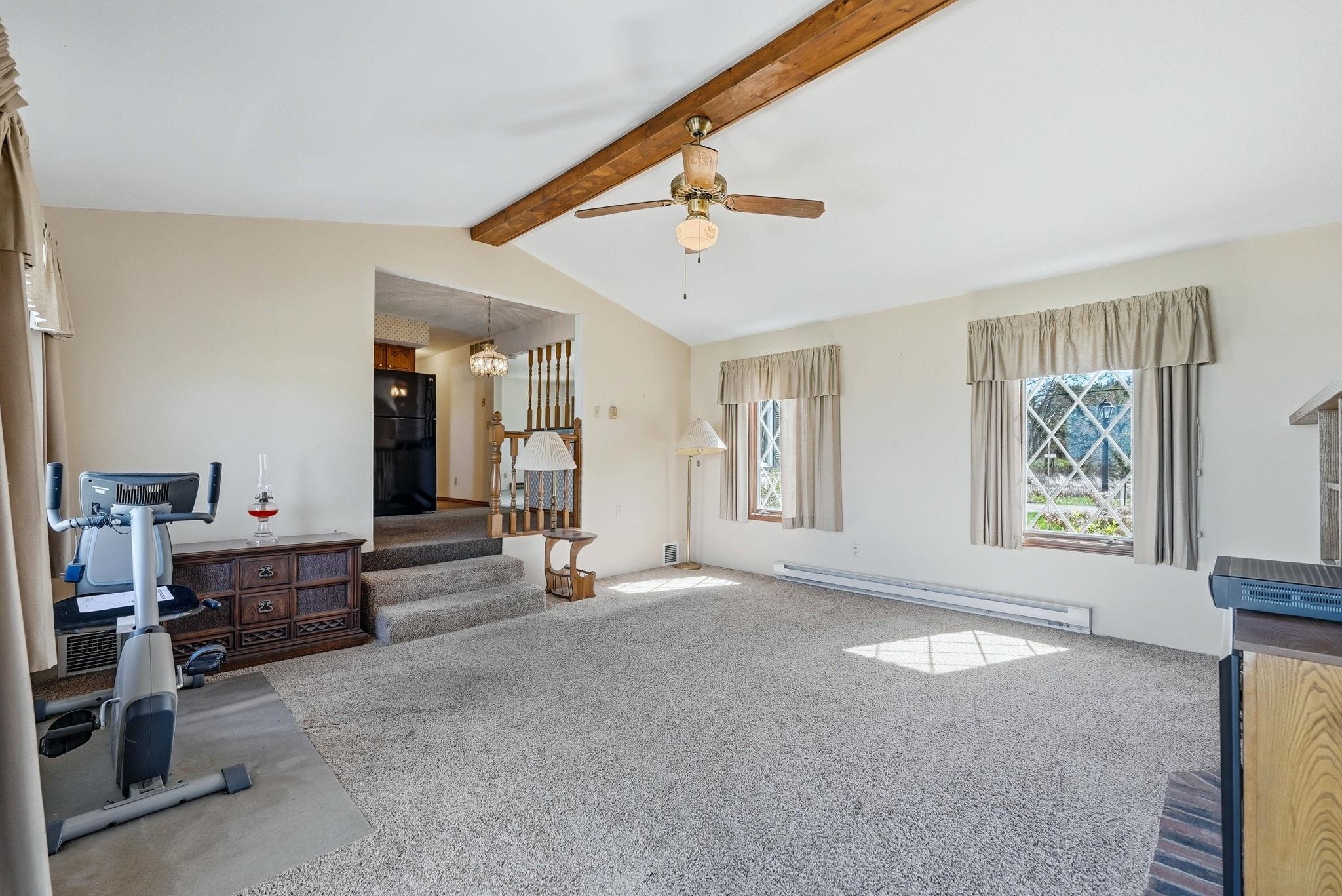Spacious living area featuring a vaulted ceiling with exposed wood beam, ceiling fan, and light-colored carpeting