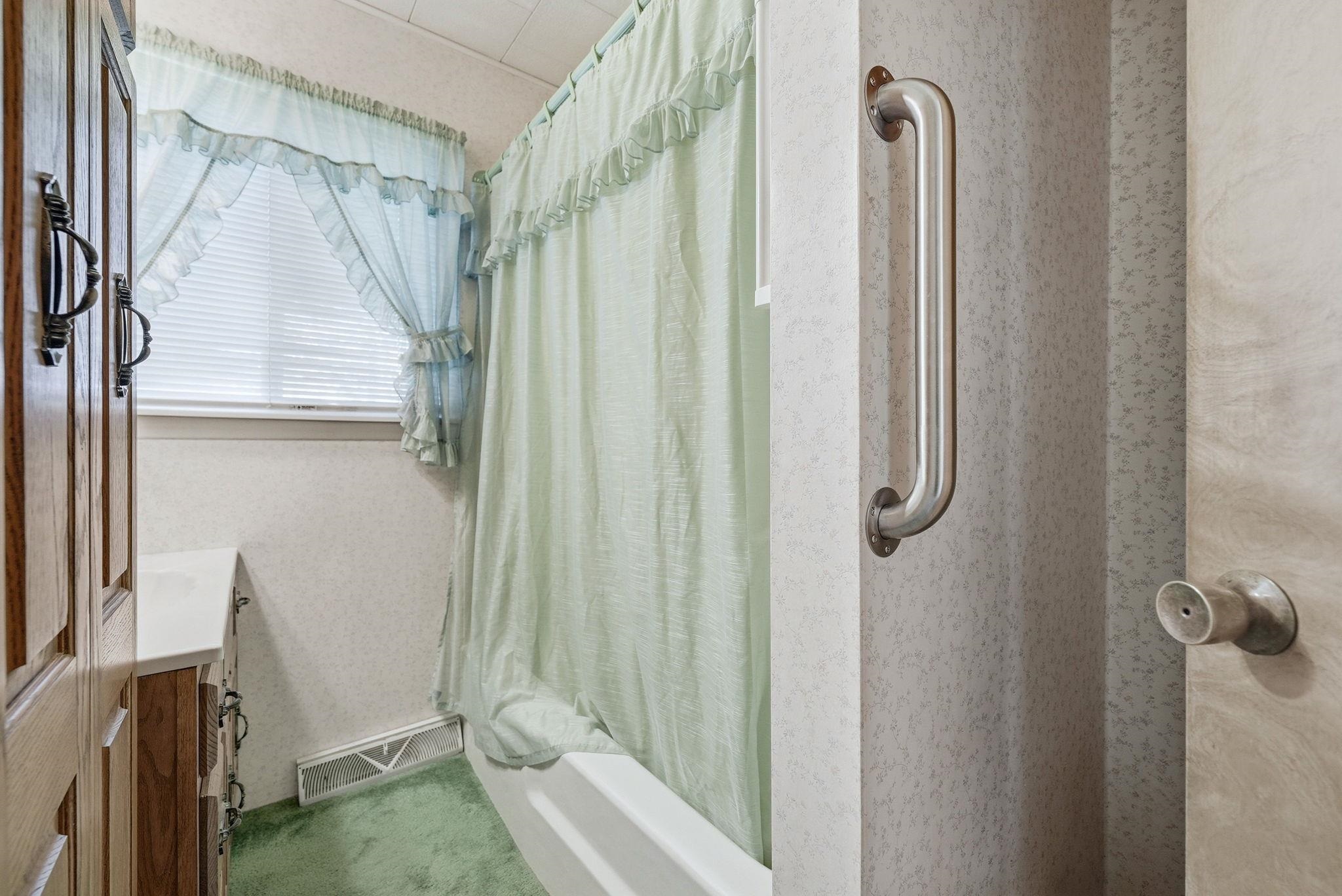 Bathroom featuring a built-in bathtub with shower curtain, window with blinds, vanity with wood-finish cabinetry, and wall-to-wall carpeting