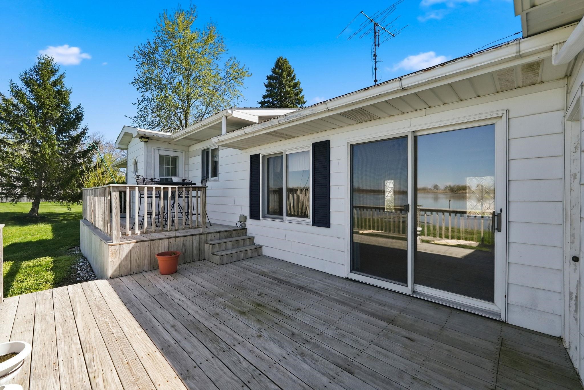 Expansive outdoor deck featuring wood flooring, a sliding glass door, white siding, a raised wooden deck with railings, and a water backdrop