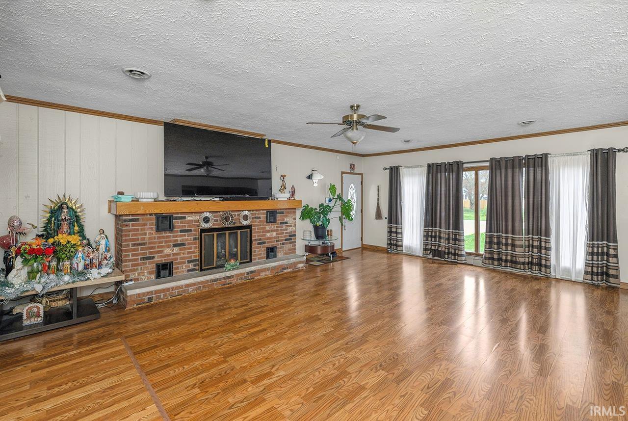 Unfurnished living room with ceiling fan, a brick fireplace, wood finished floors, a textured ceiling, and crown molding