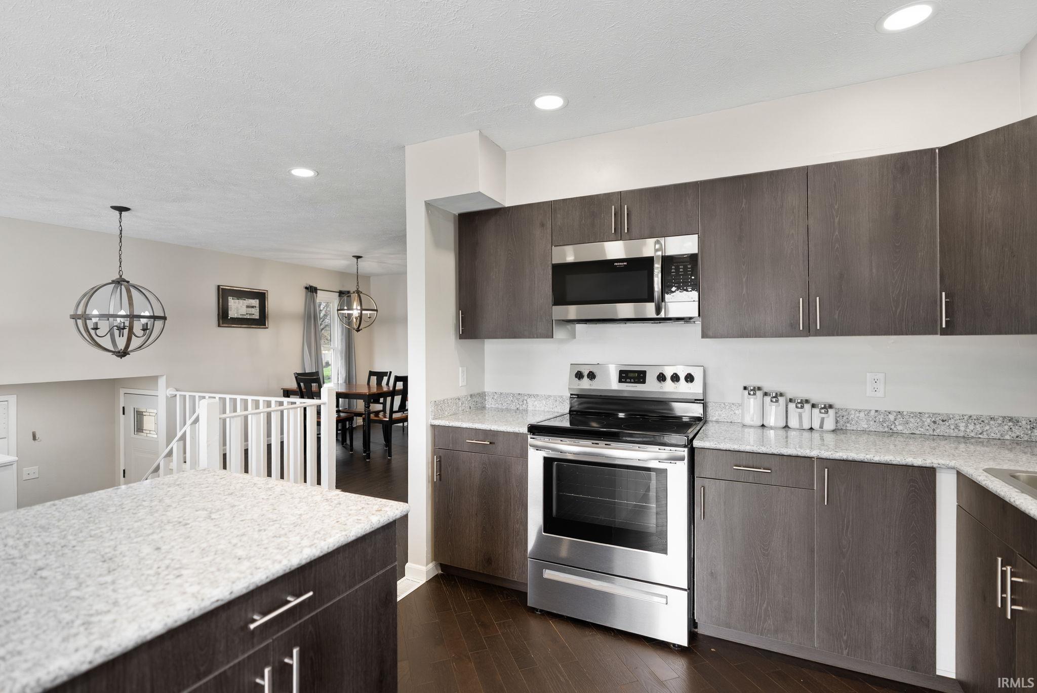 Kitchen featuring dark wood finish cabinetry, stainless steel appliances, dark wood finished floors, suspended lighting, and light stone countertops