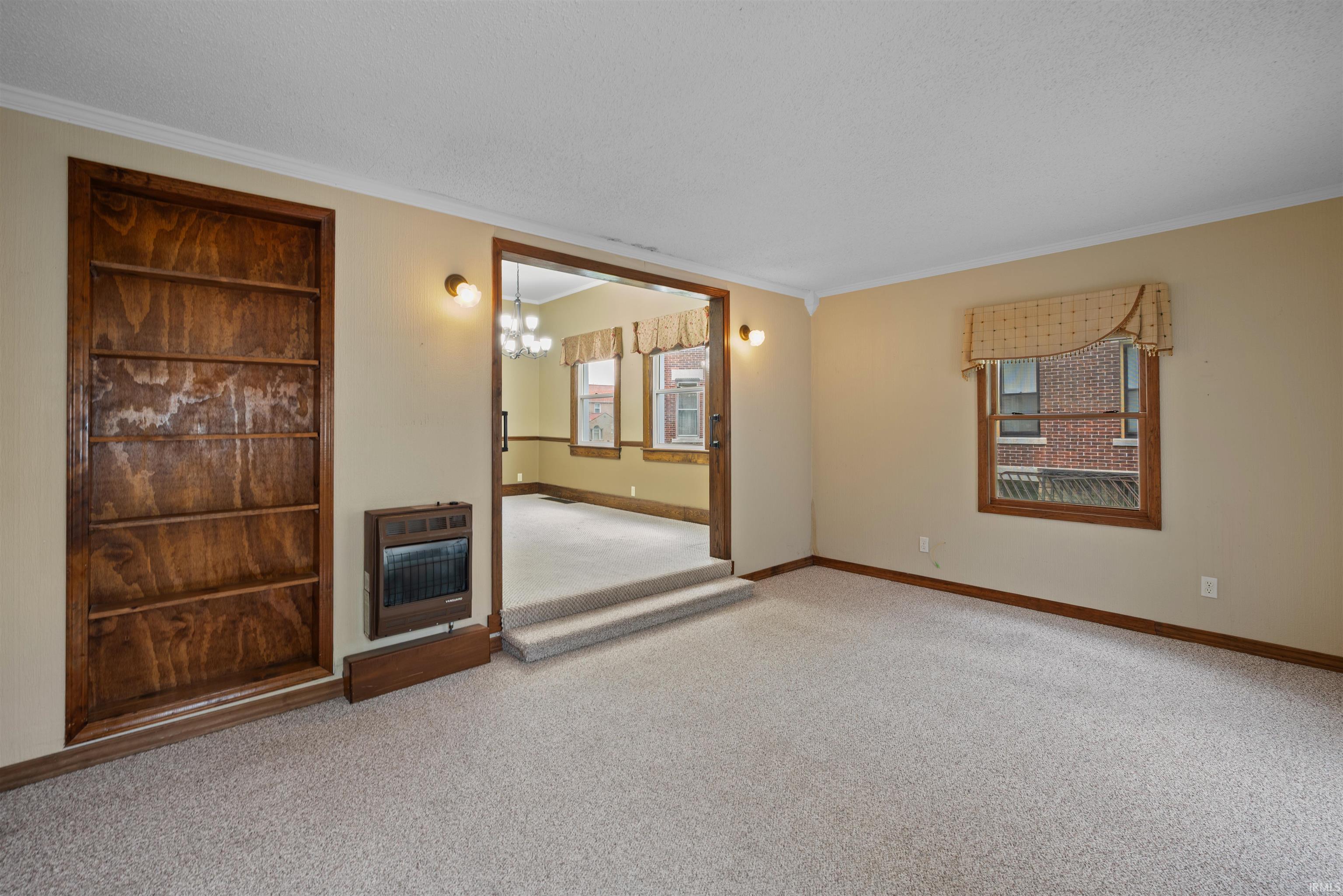 Unfurnished living room with heating unit, light colored carpet, crown molding, suspended lighting, and a textured ceiling