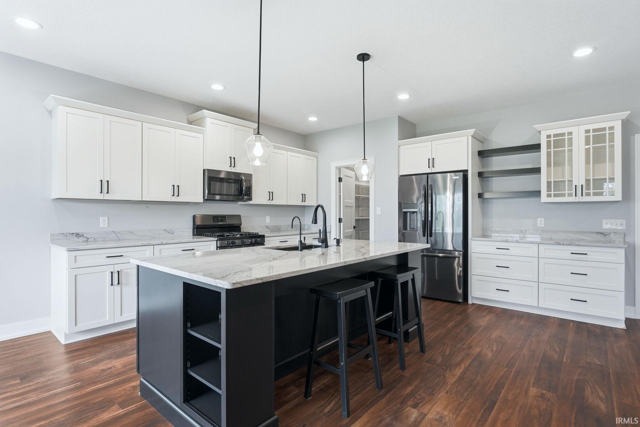 Two tone kitchen with open shelves, stainless steel appliances, light stone countertops, dark wood-type flooring, and a center island with sink