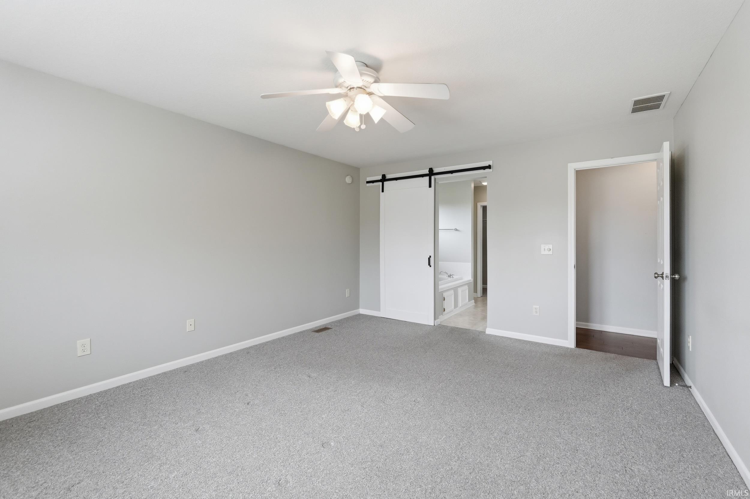 Unfurnished bedroom with a barn door, light colored carpet, ensuite bathroom, and ceiling fan