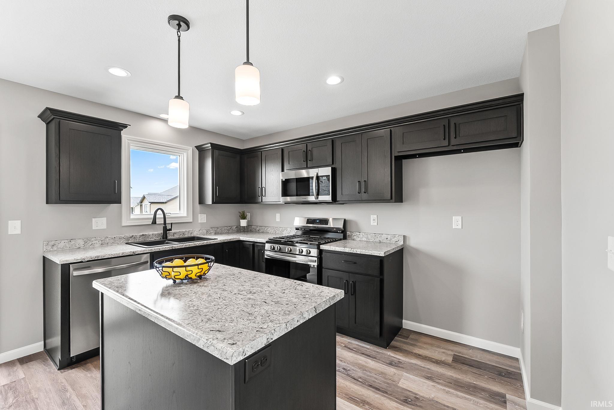 Modern kitchen featuring dark cabinetry, light-toned countertops, wood-finish flooring, stainless steel appliances, and a central island