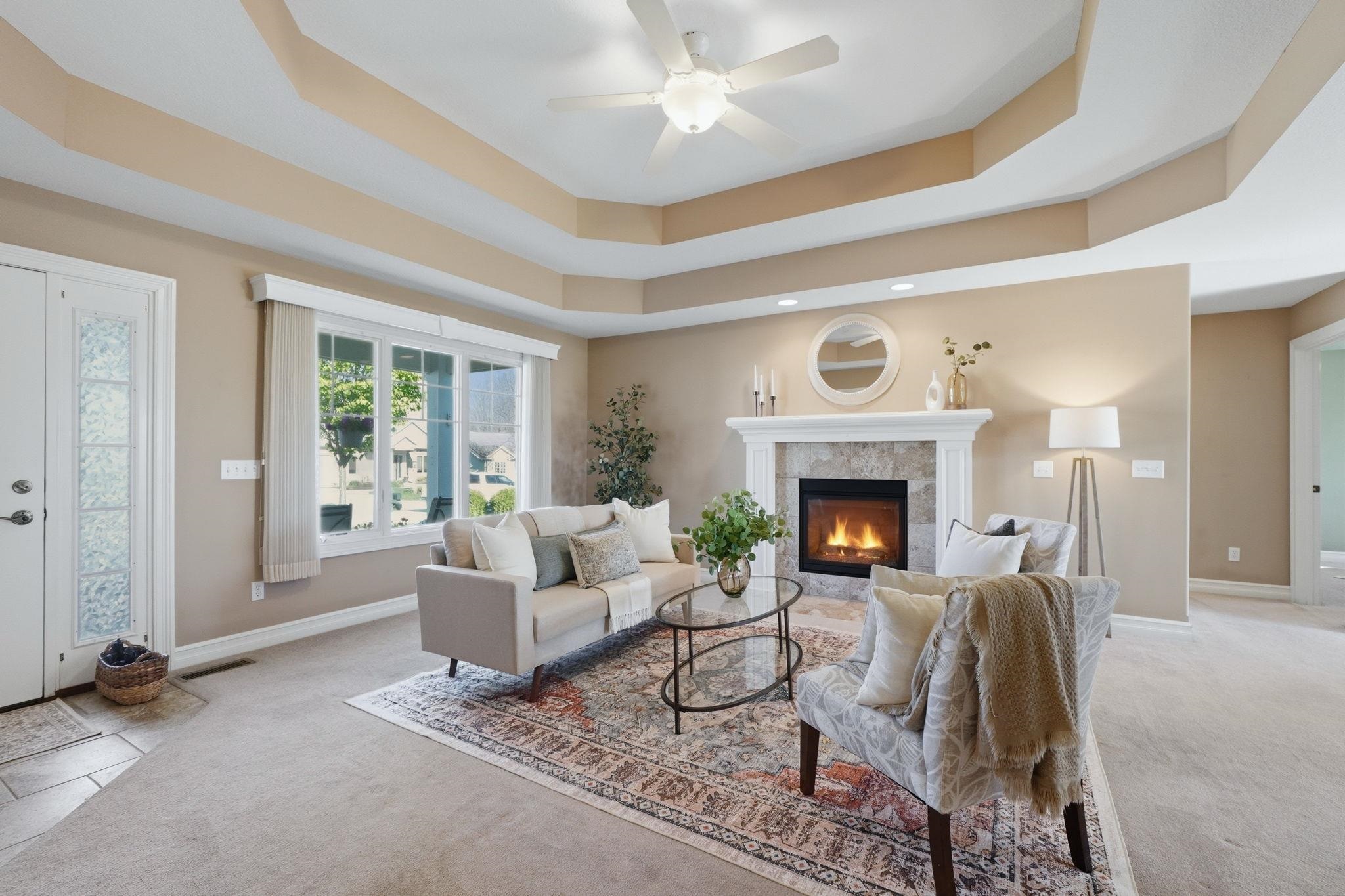 Living area with light carpet, ceiling fan, a tiled fireplace, and a raised ceiling