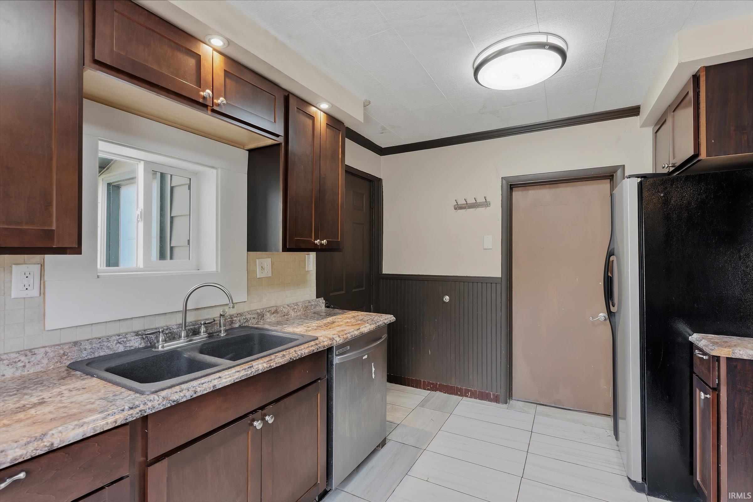 Kitchen with dark wood finish cabinetry, stainless steel appliances, a wainscoted wall, crown molding, and light stone counters