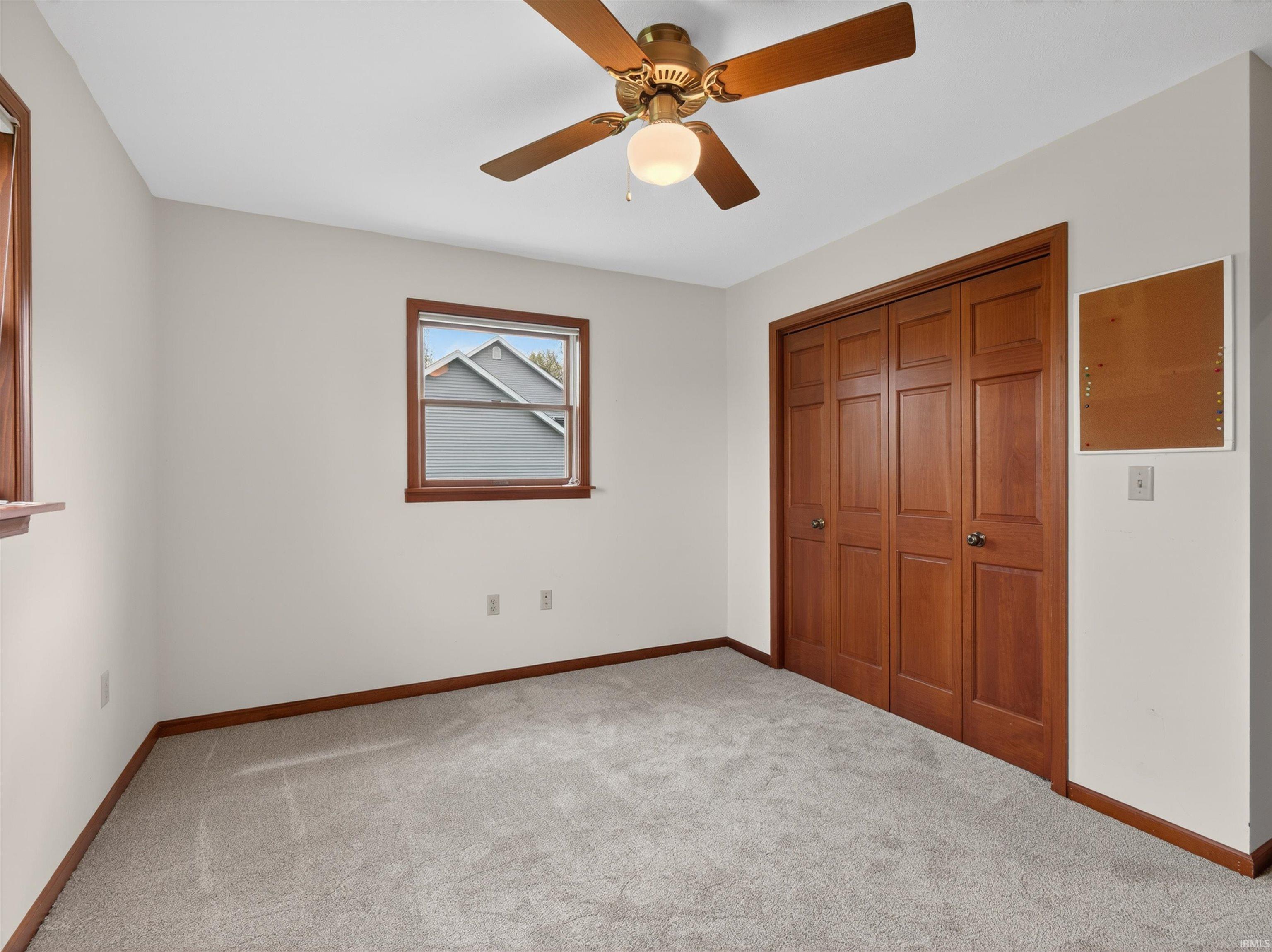 Carpeted room featuring a ceiling fan with light fixture, two windows with wood trim, and a bi-fold closet with wood panel doors