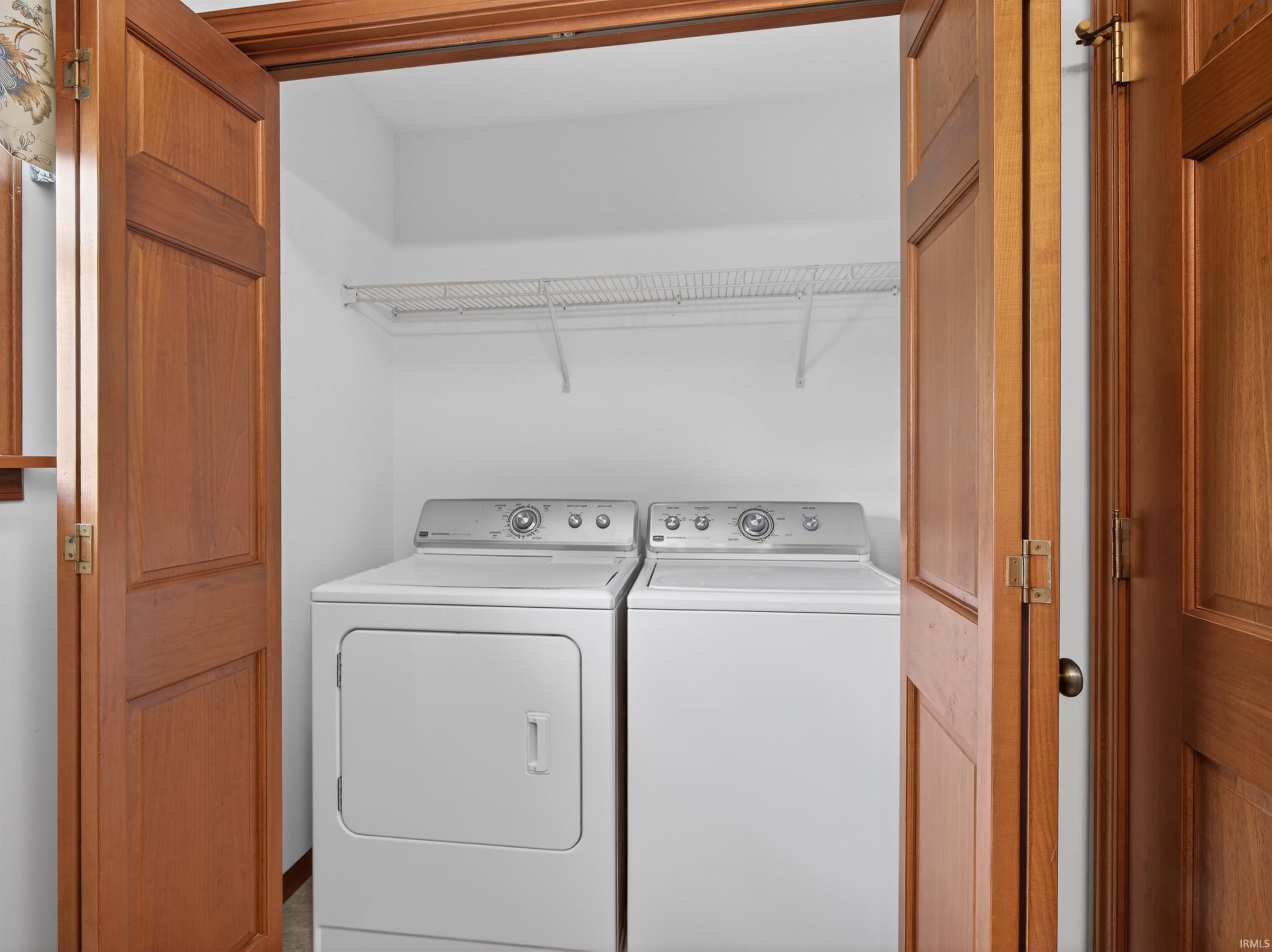 Dedicated laundry area featuring a white washer and dryer pair, an upper wire shelf for storage, and warm wood-finish bi-fold doors