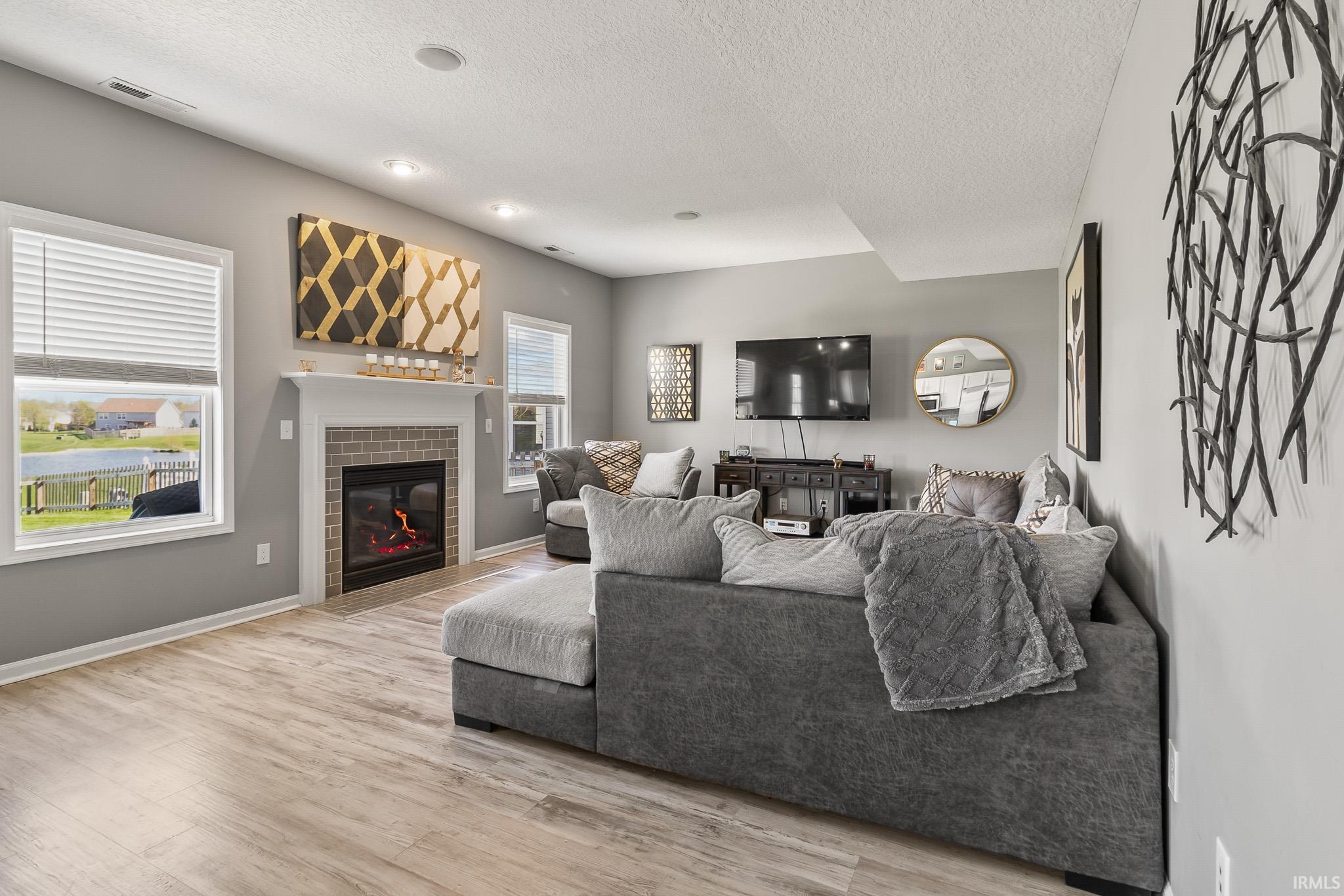Living area featuring a fireplace with a brick surround and white mantel, wood-finish flooring, recessed lighting, and multiple windows