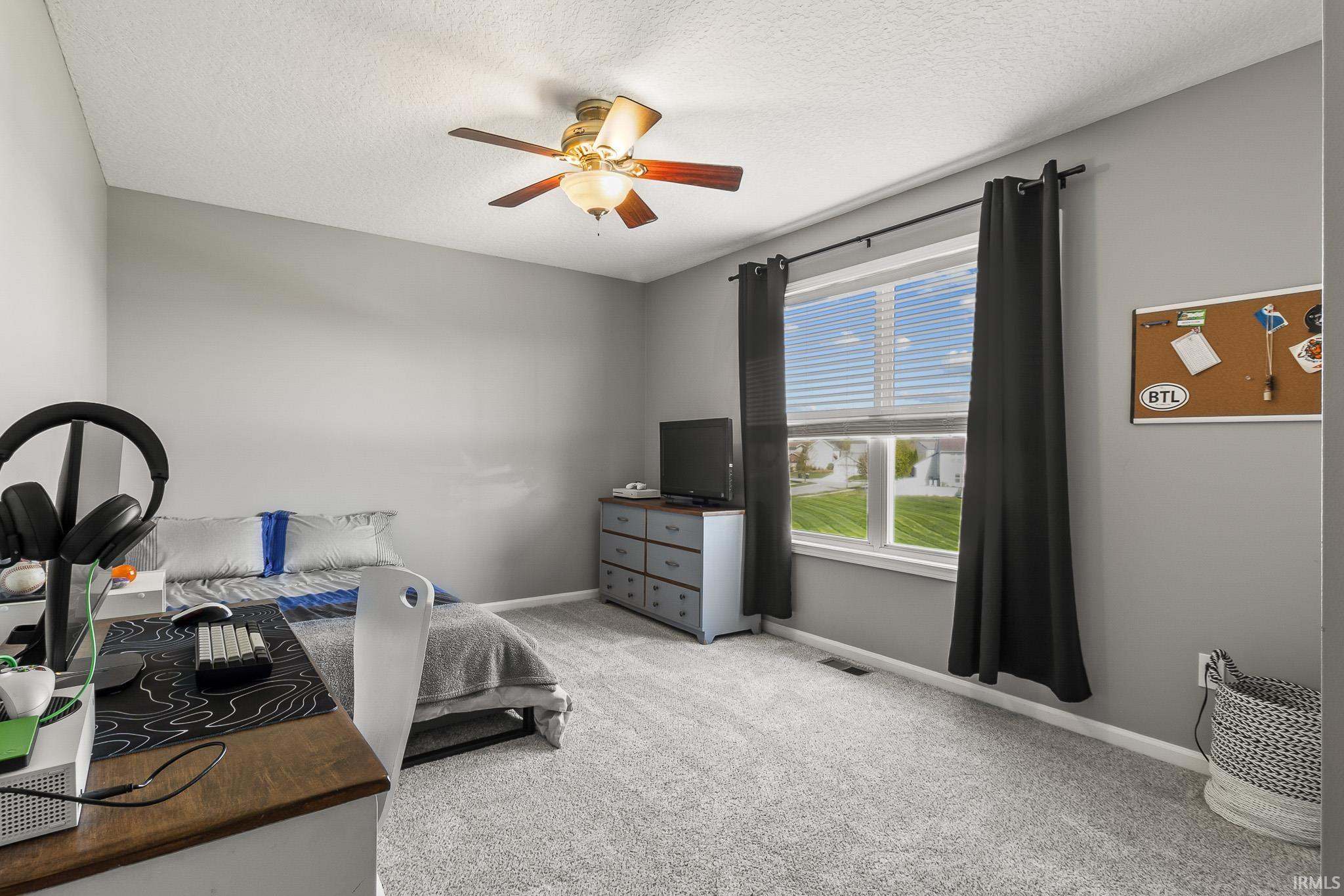 Spacious room featuring neutral grey wall paint, plush grey carpeting, a large window with white blinds, and a ceiling fan with integrated lighting