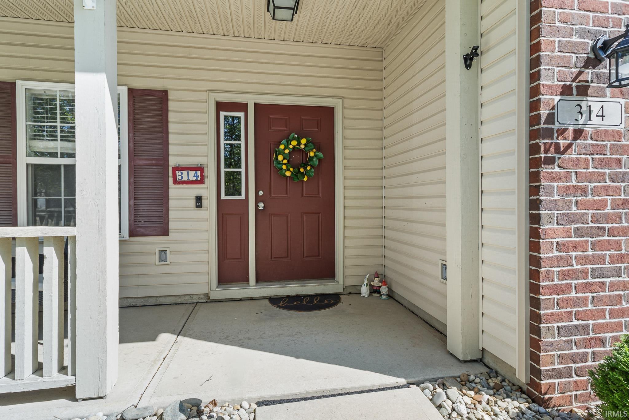 Covered front porch featuring a dark red entry door with sidelight, light-colored siding, red brick accents, and a traditional outdoor light fixture