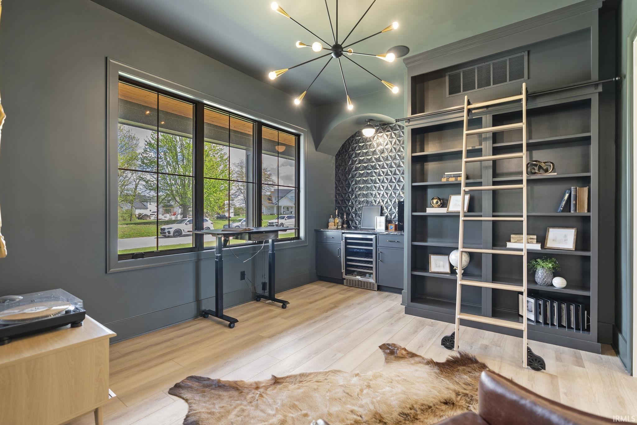 Spacious room featuring light wood-finish flooring, a large multi-pane window, built-in shelving with a rolling ladder, a wet bar with integrated beverage refrigeration, and a modern sputnik chandelier