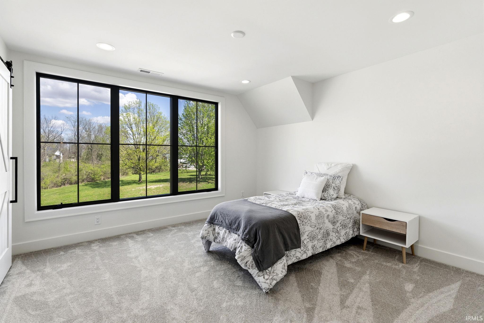 Spacious room featuring a large black-framed window with multiple panes, recessed ceiling lighting, light gray carpeting, crisp white walls, and a white barn door with a matte black handle