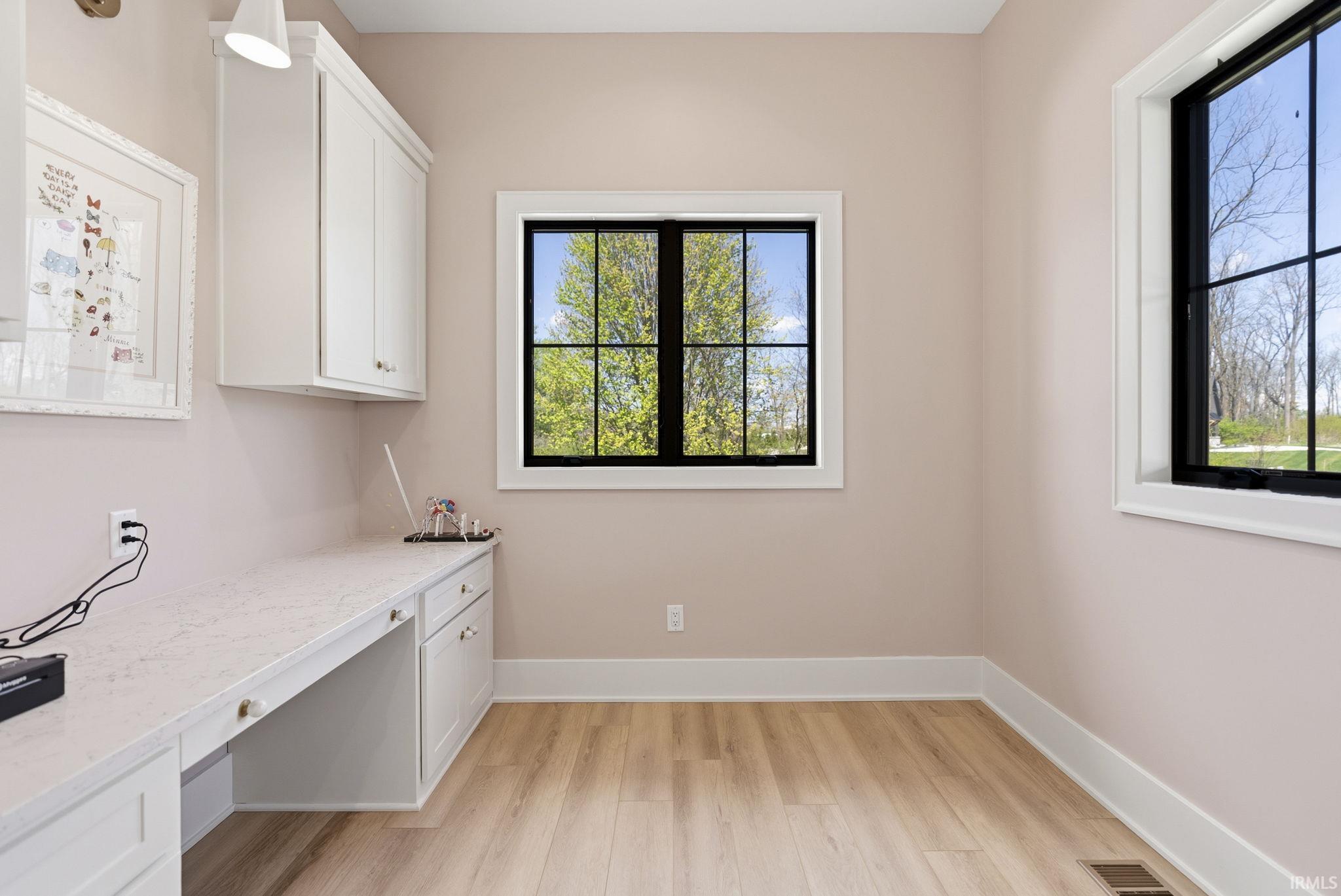 Room featuring light wood-finish flooring, two large windows with black gridded frames, and a built-in desk with white cabinetry and a light-toned countertop