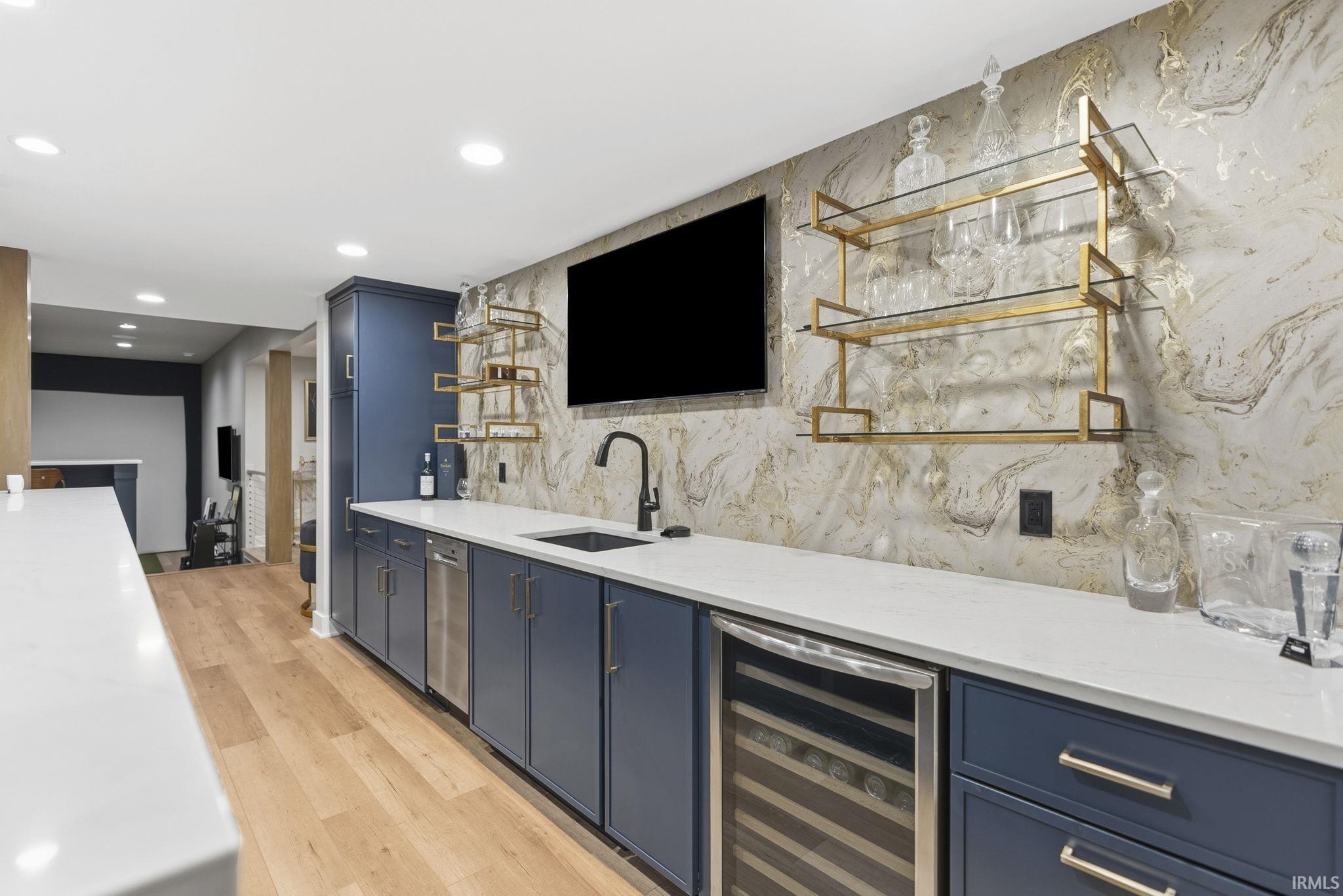 Wet bar featuring custom blue cabinetry, white countertops, a stainless steel beverage refrigerator, a built-in sink with a matte black faucet, and decorative gold-toned shelving against a patterned wall