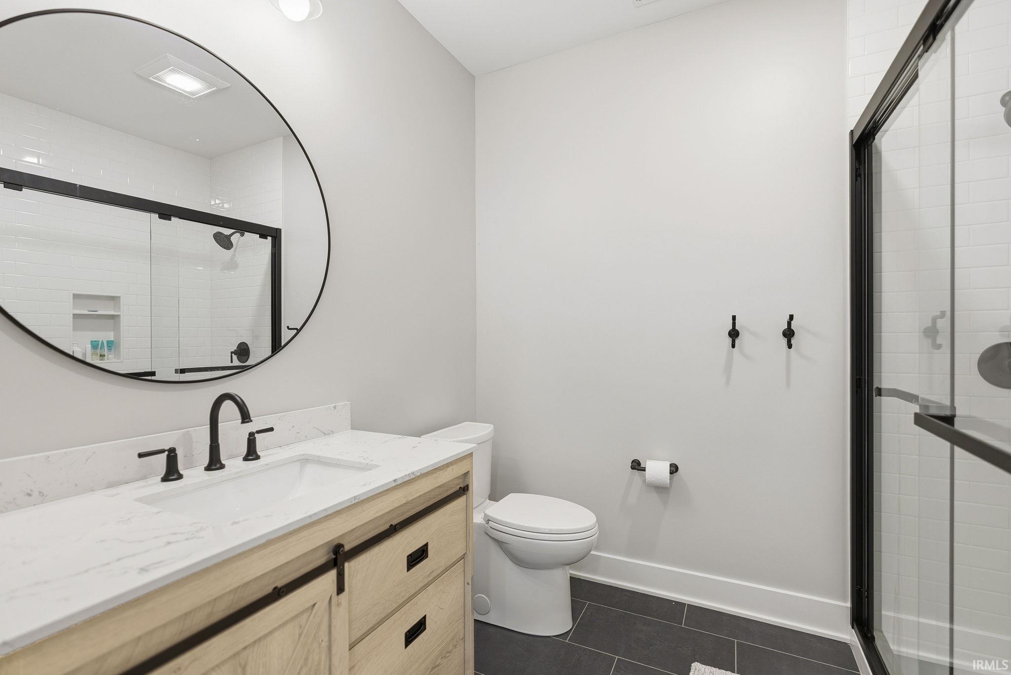 Bathroom featuring a light wood vanity with a white countertop and undermount sink, a matte black faucet, a circular mirror, and a glass-enclosed shower with black framing