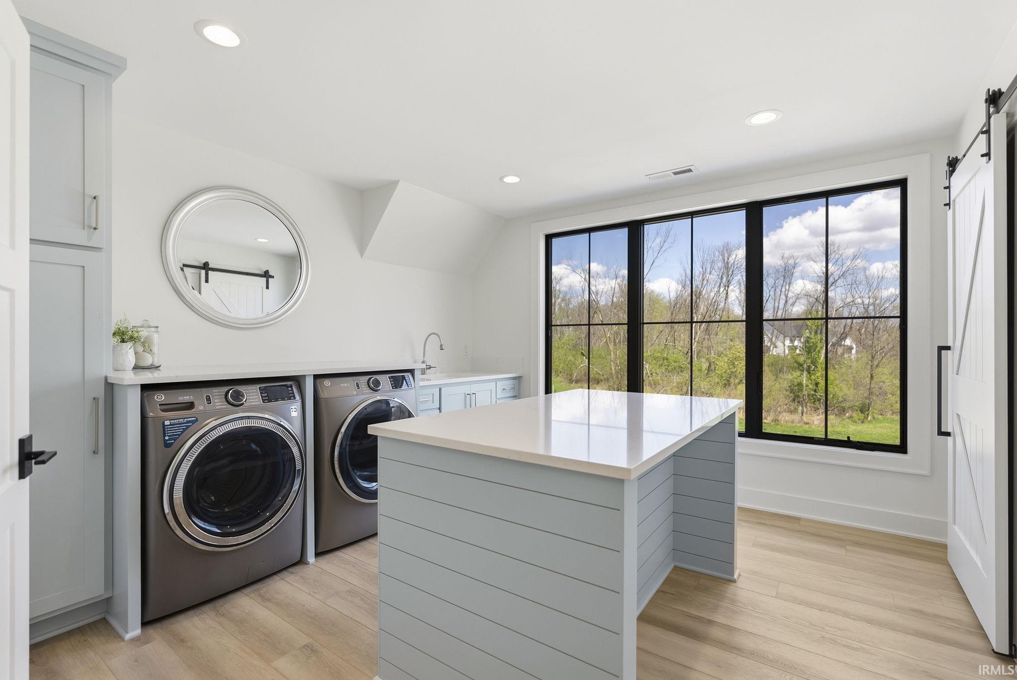Spacious utility room featuring a light wood-finish floor, a large window with black framing, recessed lighting, light blue built-in cabinetry, and a central island with a light-toned countertop