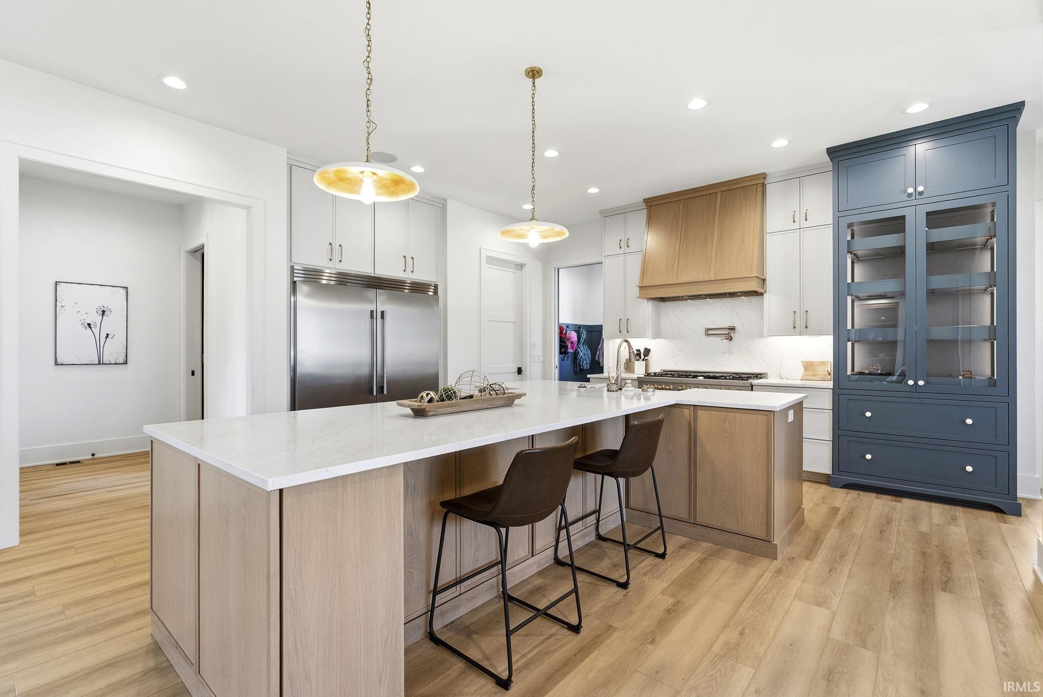 Spacious kitchen featuring a large island with a light-toned wood-finish base and a light countertop, stainless steel appliances, a range with a wood-clad hood, light cabinetry, and a prominent blue built-in hutch with glass-front uppers