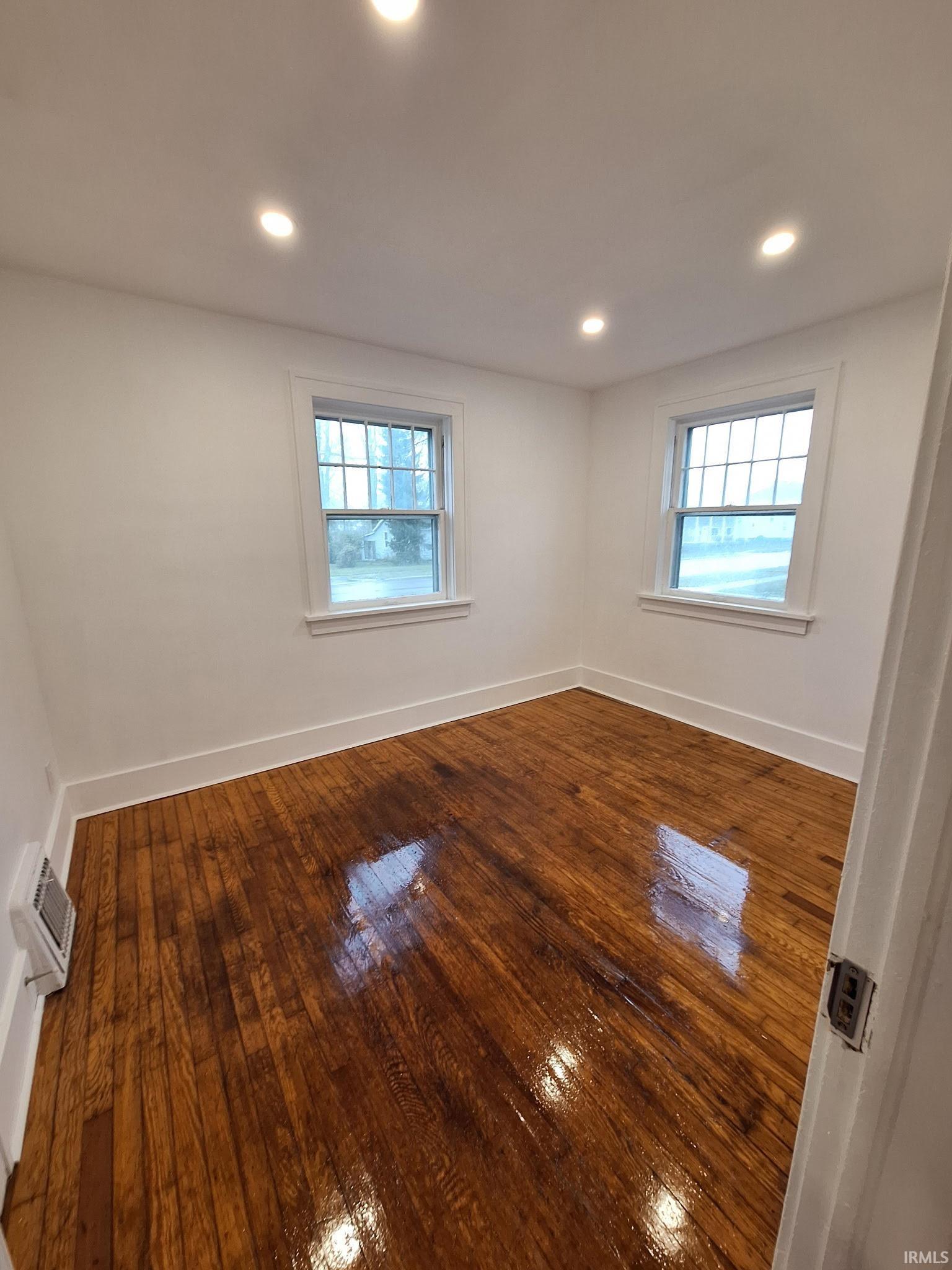 Room featuring rich wood flooring, white walls, two windows with white trim, recessed ceiling lighting, and a baseboard heater