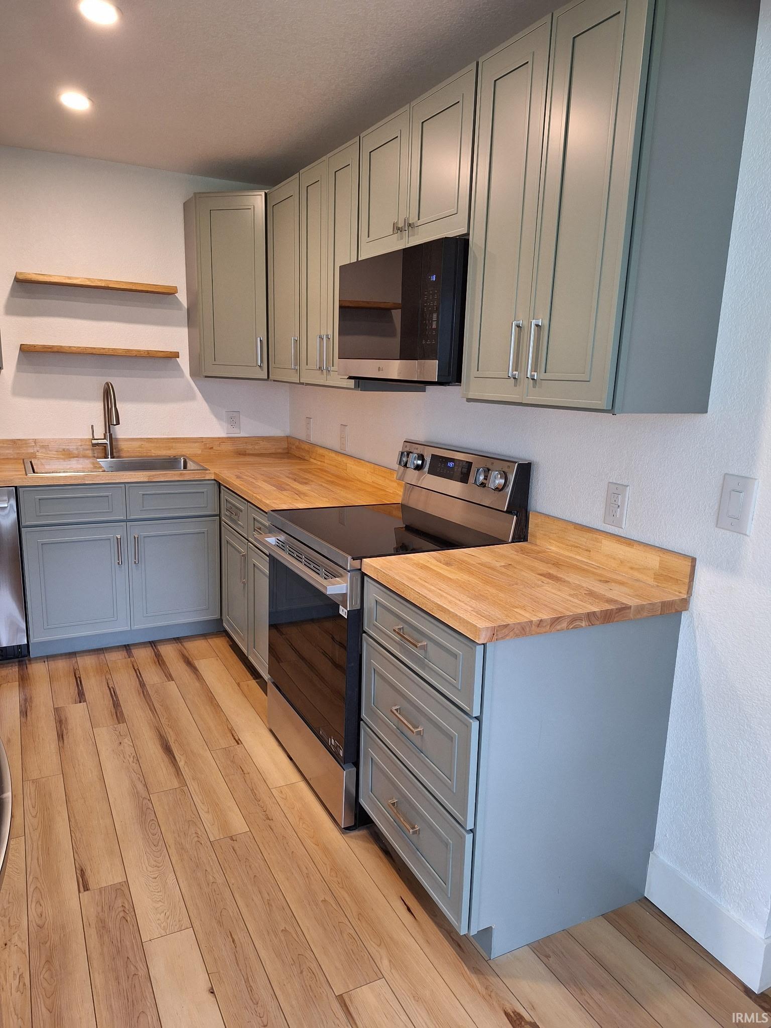 Kitchen featuring light-toned wood-finish flooring, light blue-grey cabinetry with contemporary hardware, wood countertops, a stainless steel sink with a gooseneck faucet, and a stainless steel oven/range with an overhead microwave