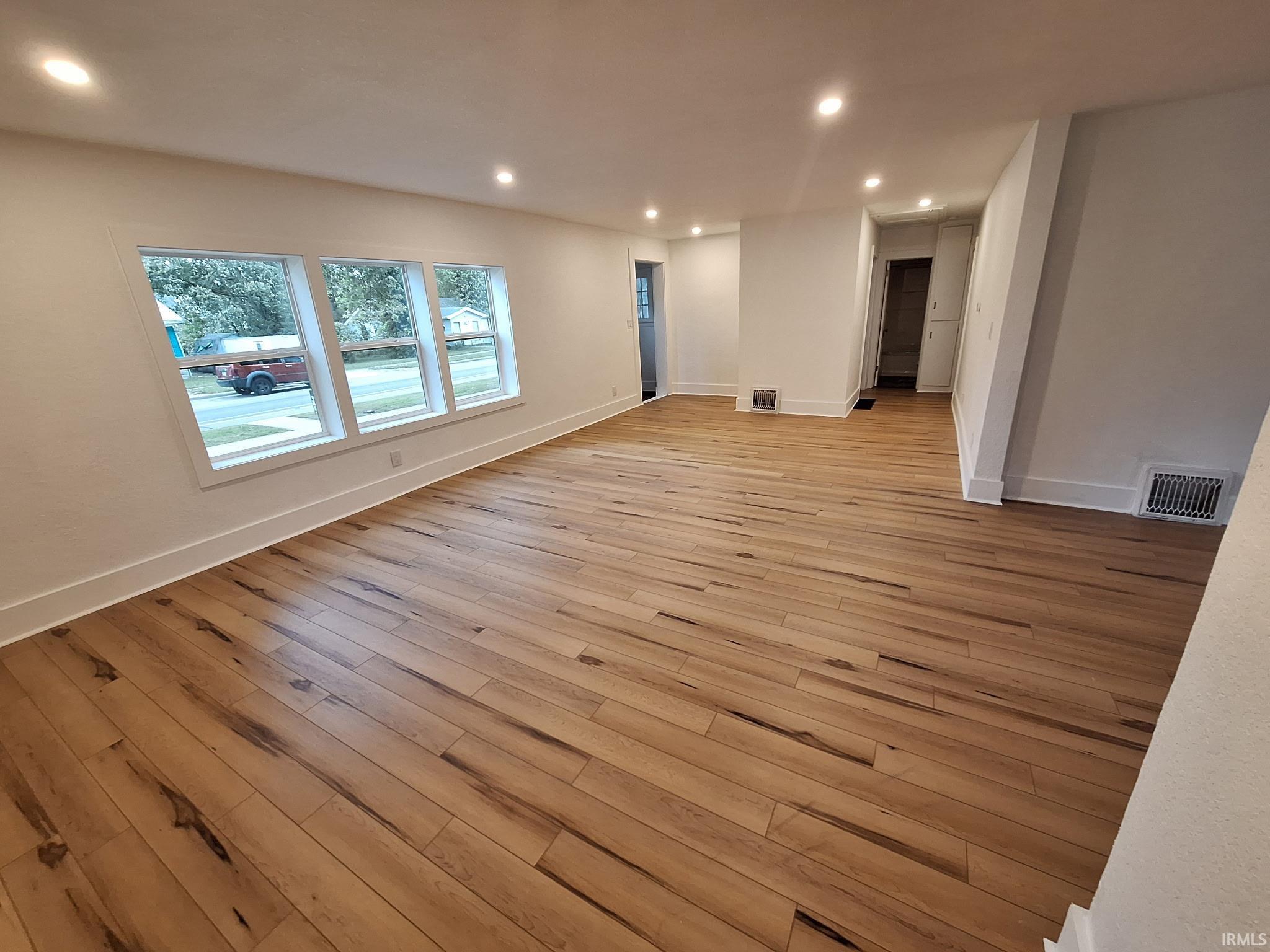Spacious living area featuring contemporary light wood-finish flooring, recessed ceiling lighting, three large windows with white trim, white baseboards, and white painted walls