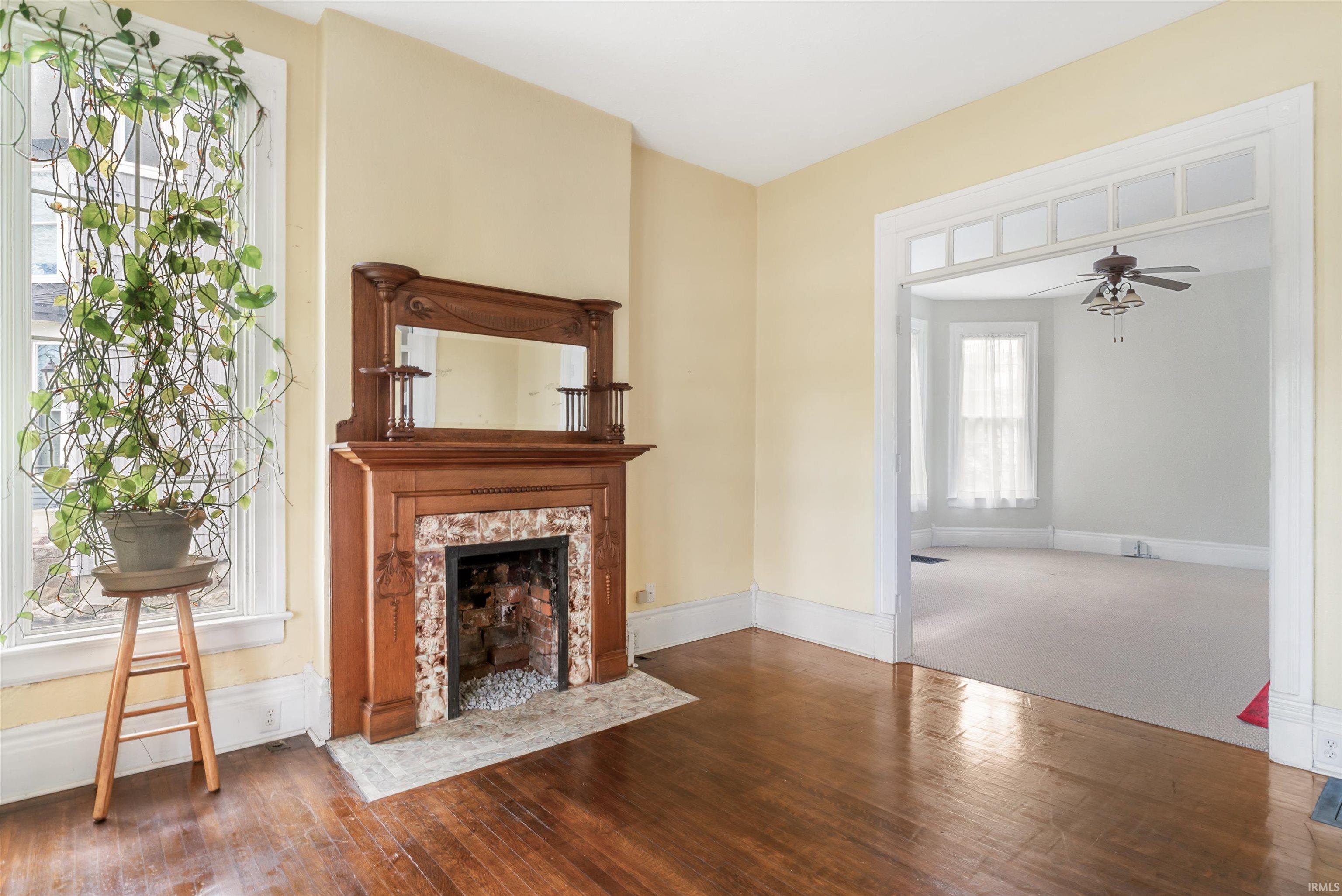 Hardwood flooring, decorative fireplace with brick and stone surround, ornate wood mantel with integrated mirror, transom window feature, and ceiling fan in an adjoining room