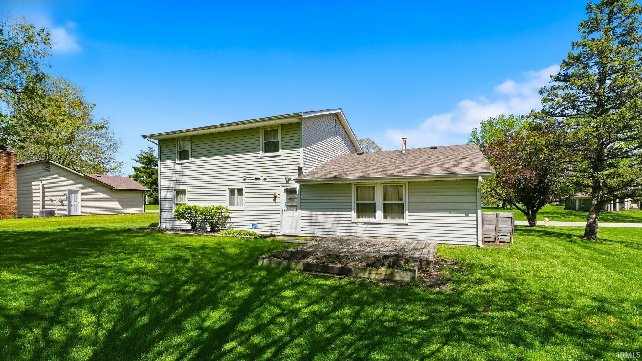 Expansive green lawn with mature trees and a two-story residence featuring light-colored siding and a multi-level patio