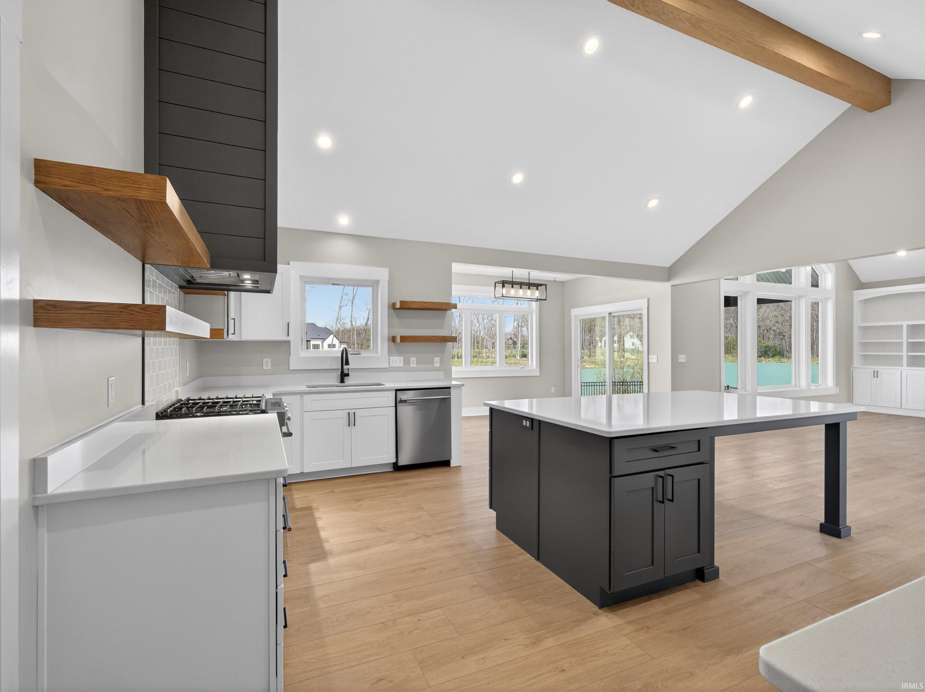 Open-concept kitchen featuring a charcoal range hood, natural wood-finish floating shelves, white cabinetry, and a dark-toned island with an integrated breakfast bar