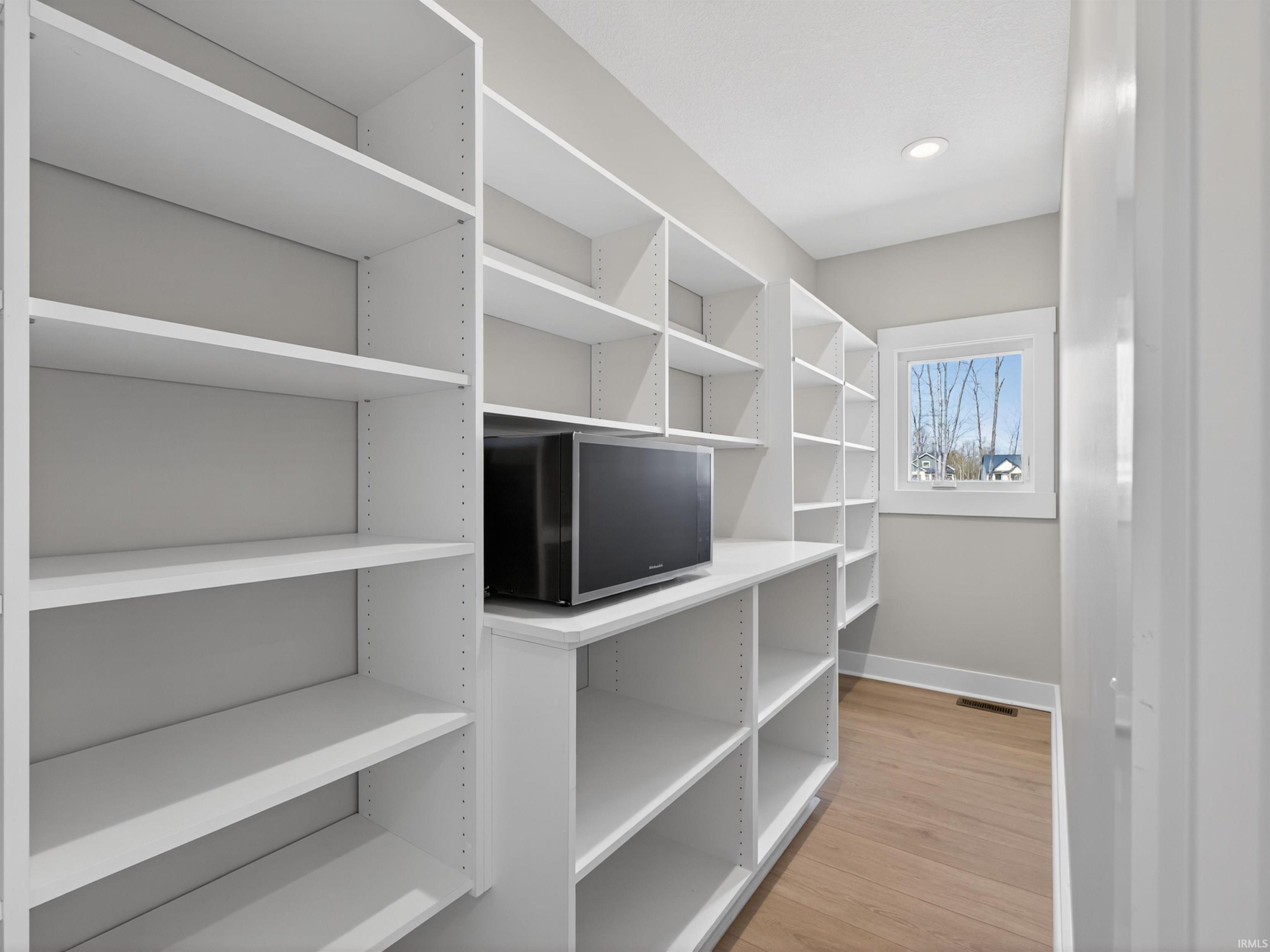 Spacious pantry featuring extensive built-in white shelving, wood-finish flooring, recessed lighting, and a single window with white trim