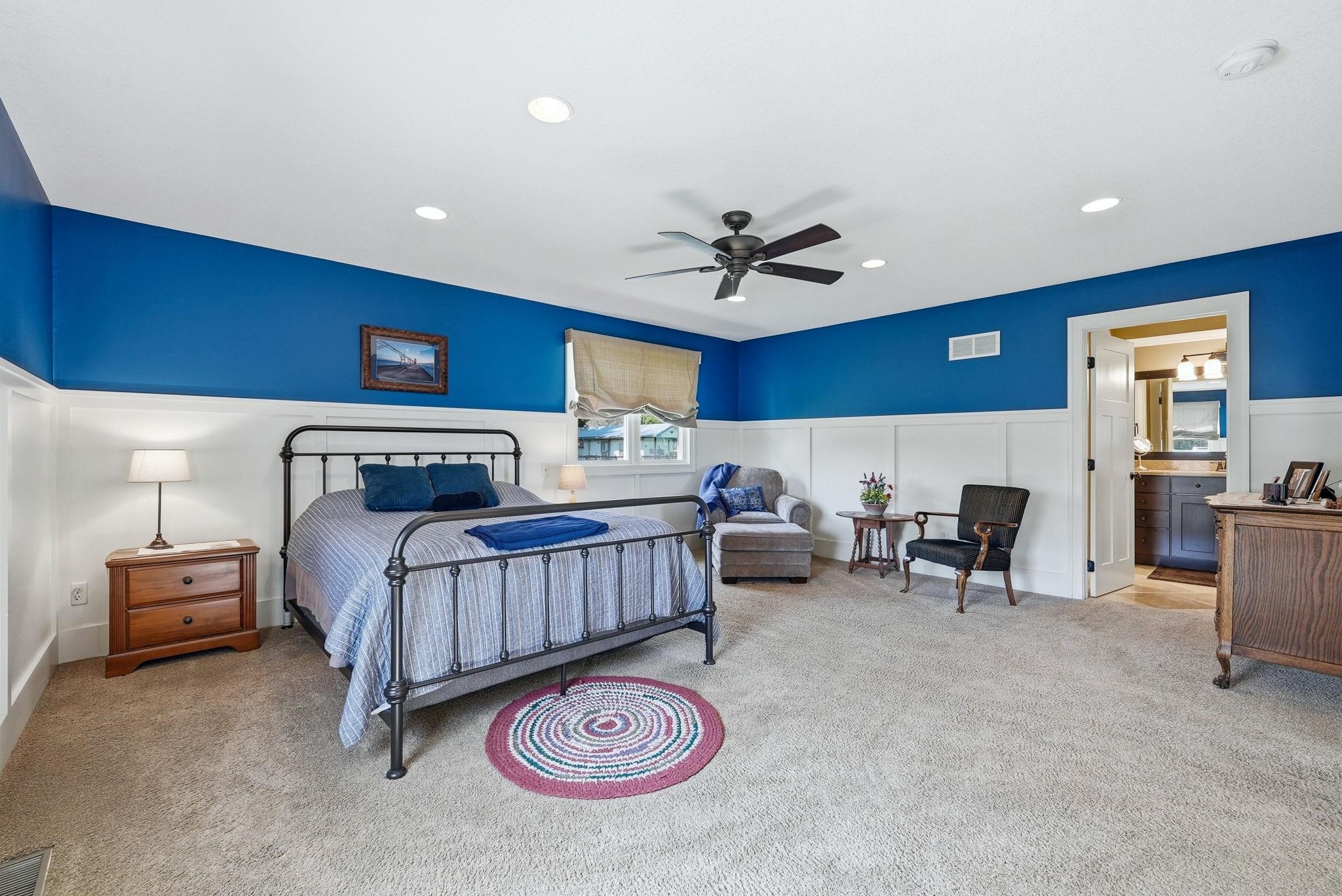 Spacious interior featuring a ceiling fan, recessed lighting, carpet flooring, wainscoting, and an open doorway to an ensuite bath with a dark wood vanity